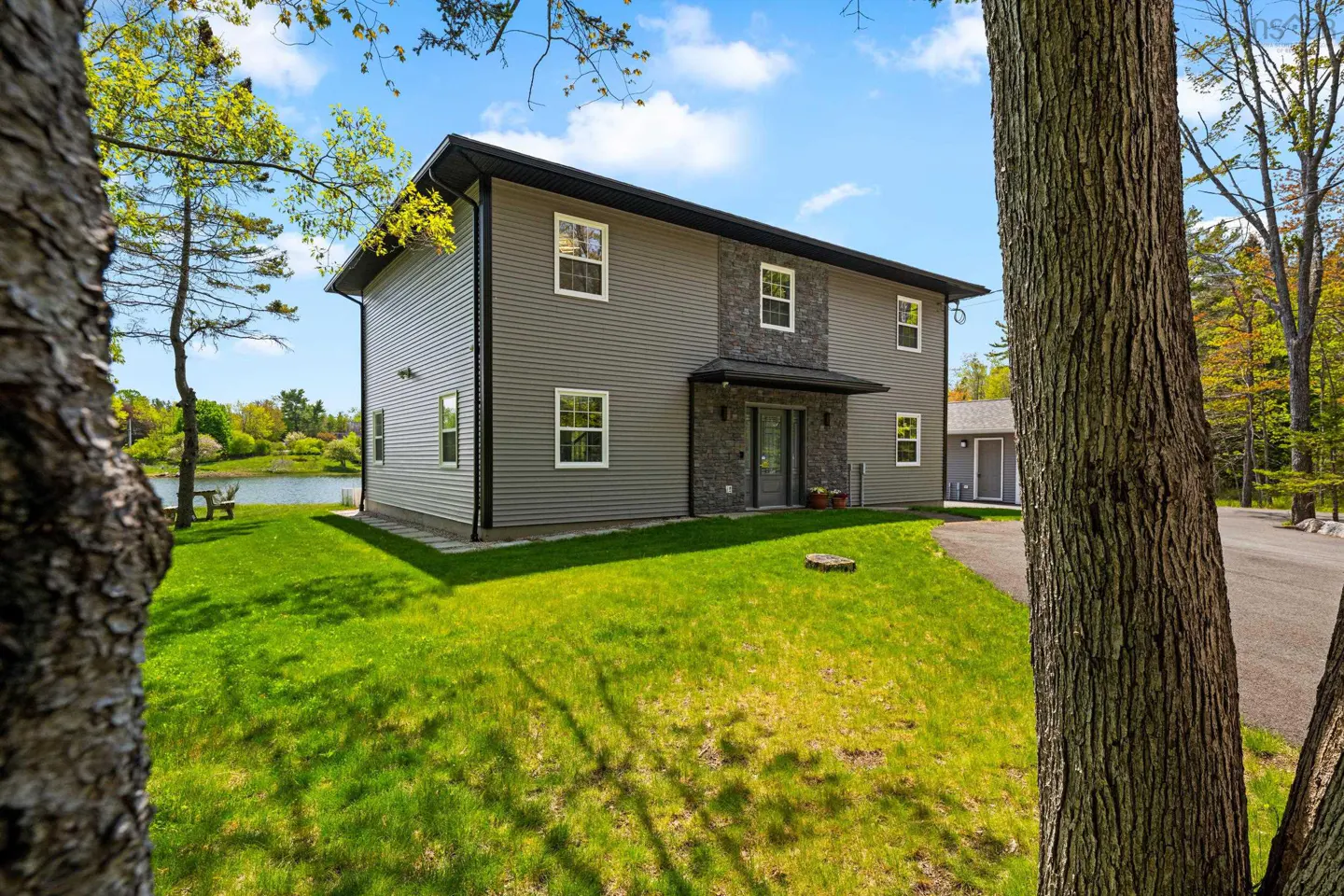 Two-story grey house with stone accents, green lawn, and lake view, framed by trees under a blue sky.