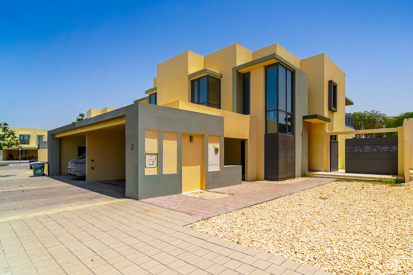 Exterior of a modern, two-story, yellow house with gray accents, a carport, and a gravel front yard.
