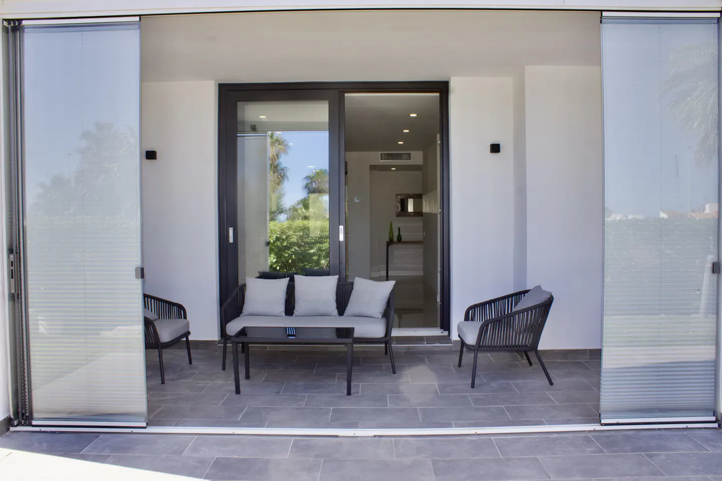 Outdoor patio with sliding glass doors, gray tile floor, black wicker furniture, and view of greenery.