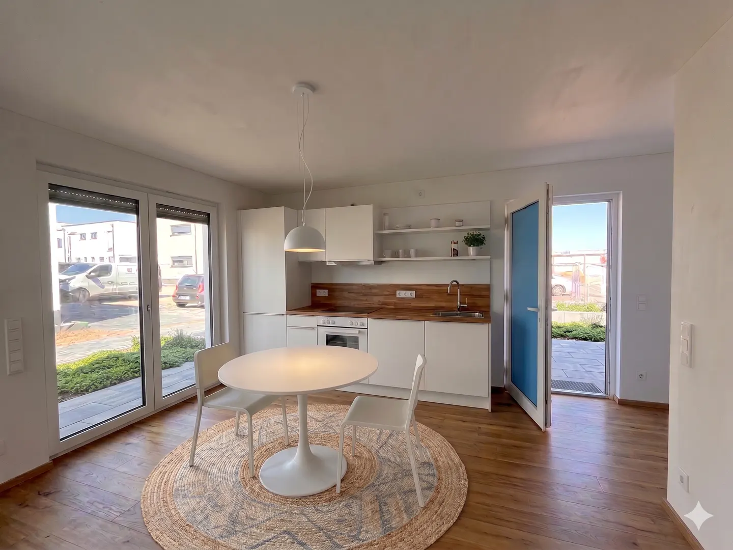 Bright, modern kitchen with white cabinets, wood countertops, and a round white table with two chairs on a jute rug. Patio doors lead to an outdoor area.