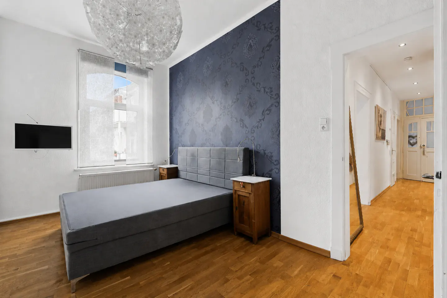 Bedroom with gray bed, wood floor, and blue accent wall. A hallway is visible through a doorway.