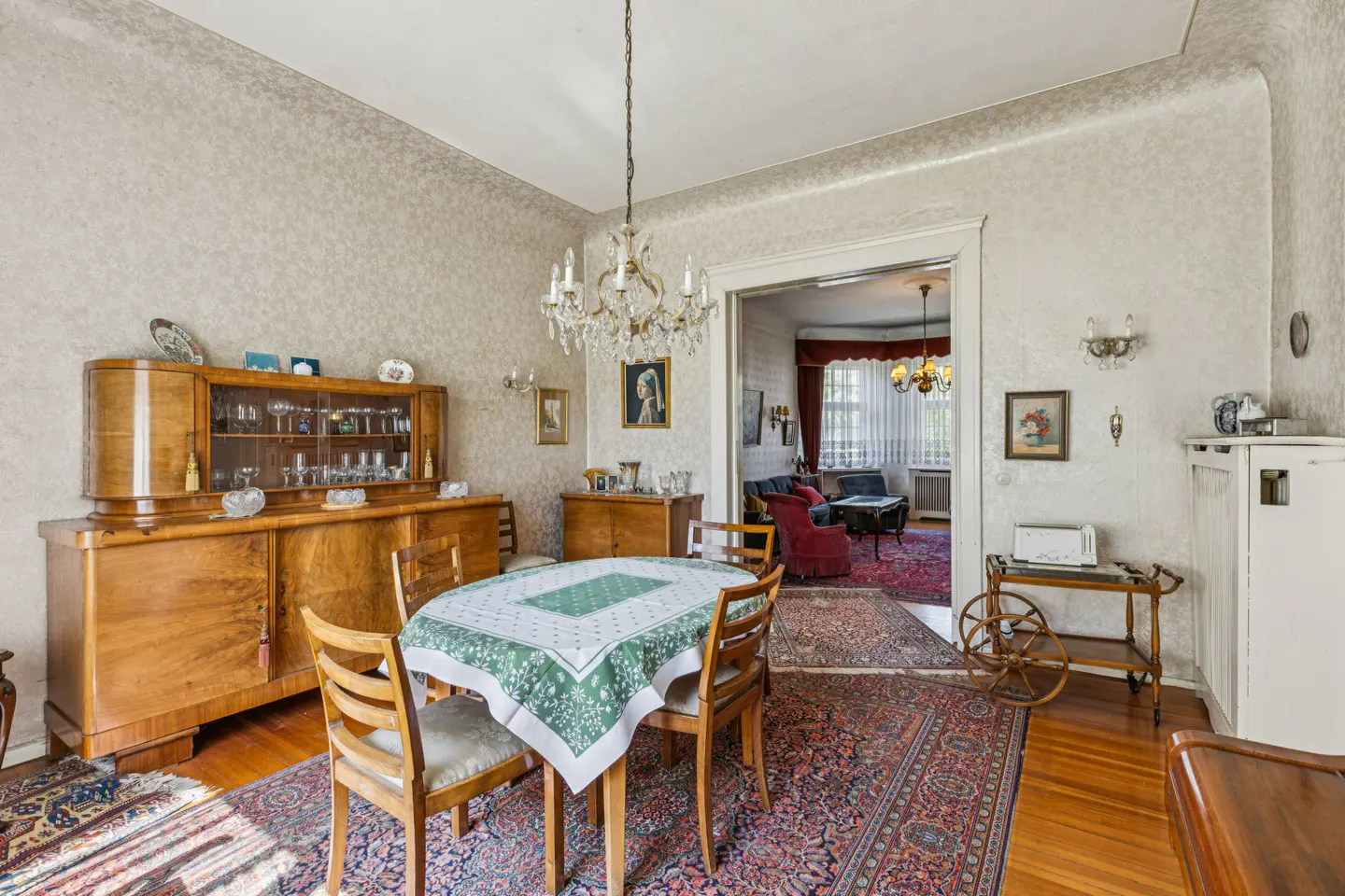 A dining room with a wooden table, chairs, and a green tablecloth. A chandelier hangs above, and a wooden cabinet stands against the patterned wallpaper.