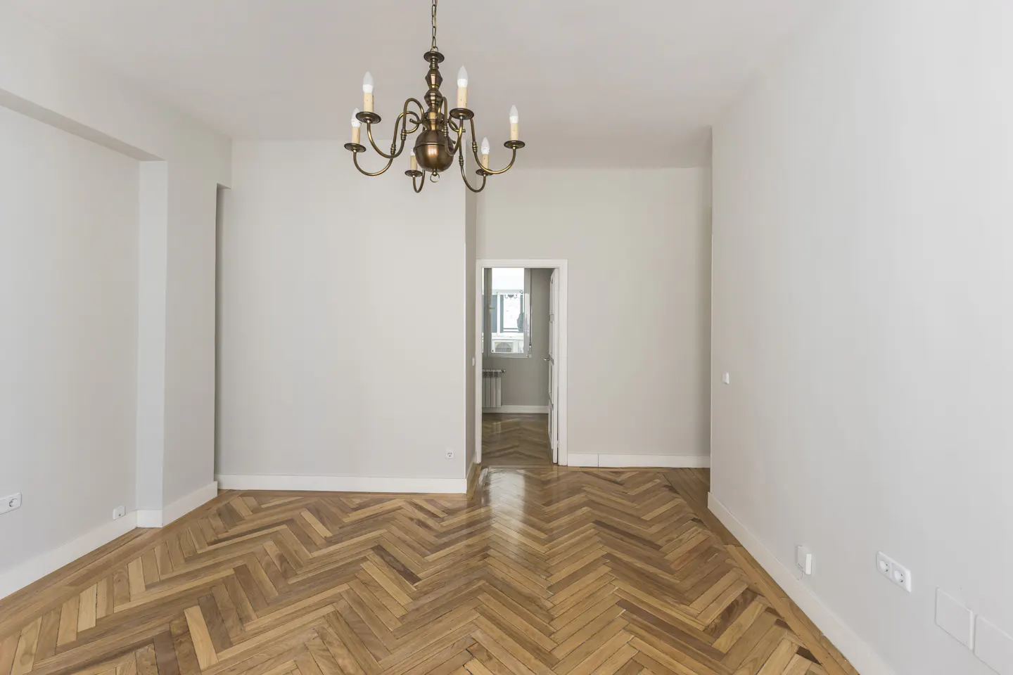 Empty room with light gray walls, polished herringbone wood floors, and a classic brass chandelier hanging from the ceiling.