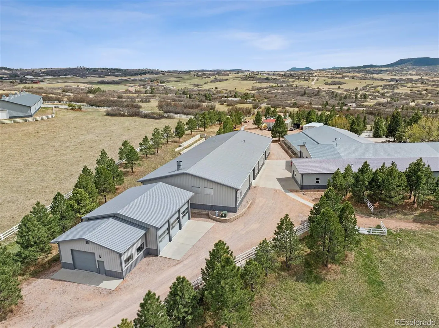 Aerial view of a large grey metal workshop and storage complex on a rural property with pine trees and rolling hills.