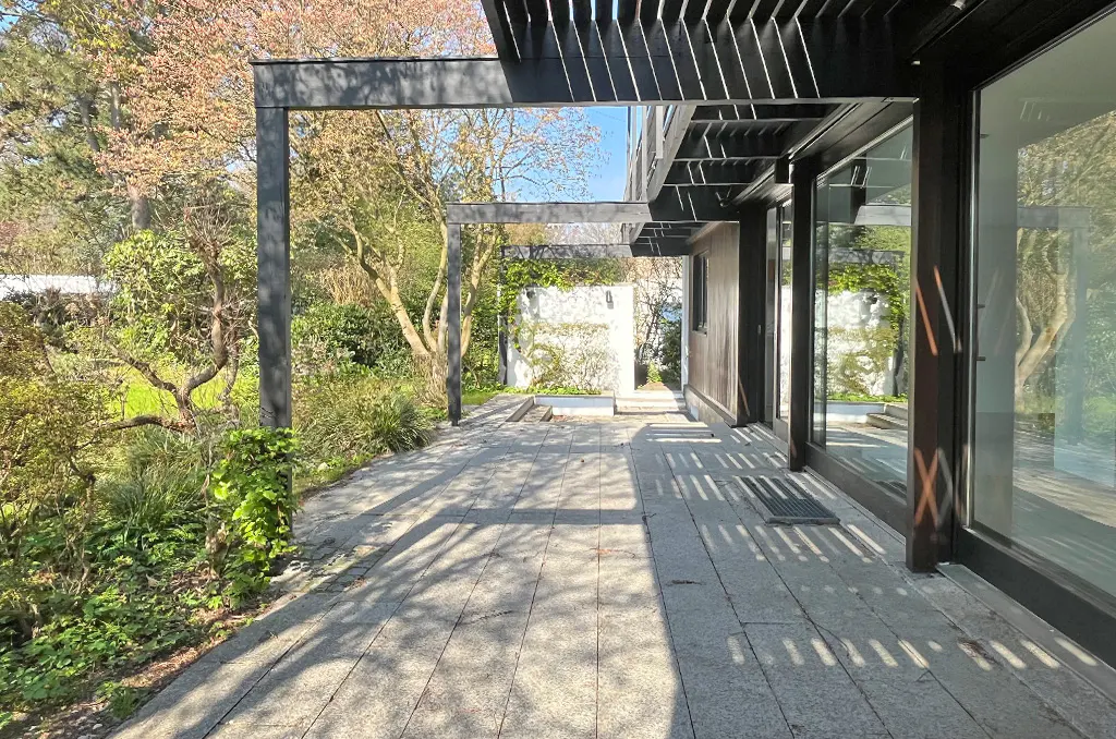 Exterior view of a modern home with a stone patio, black pergola, and lush green landscaping. Sunlight casts shadows on the patio.