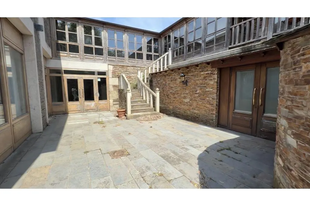 Stone-paved courtyard with a stone staircase leading to a wooden balcony, surrounded by rustic stone walls and glass doors.