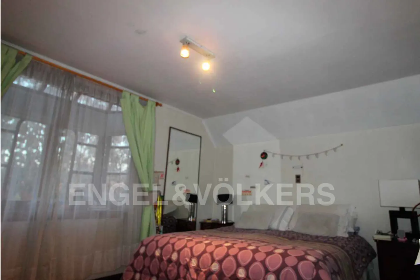 Bedroom with a bed covered in a purple patterned quilt, white walls, and a window with sheer white curtains.