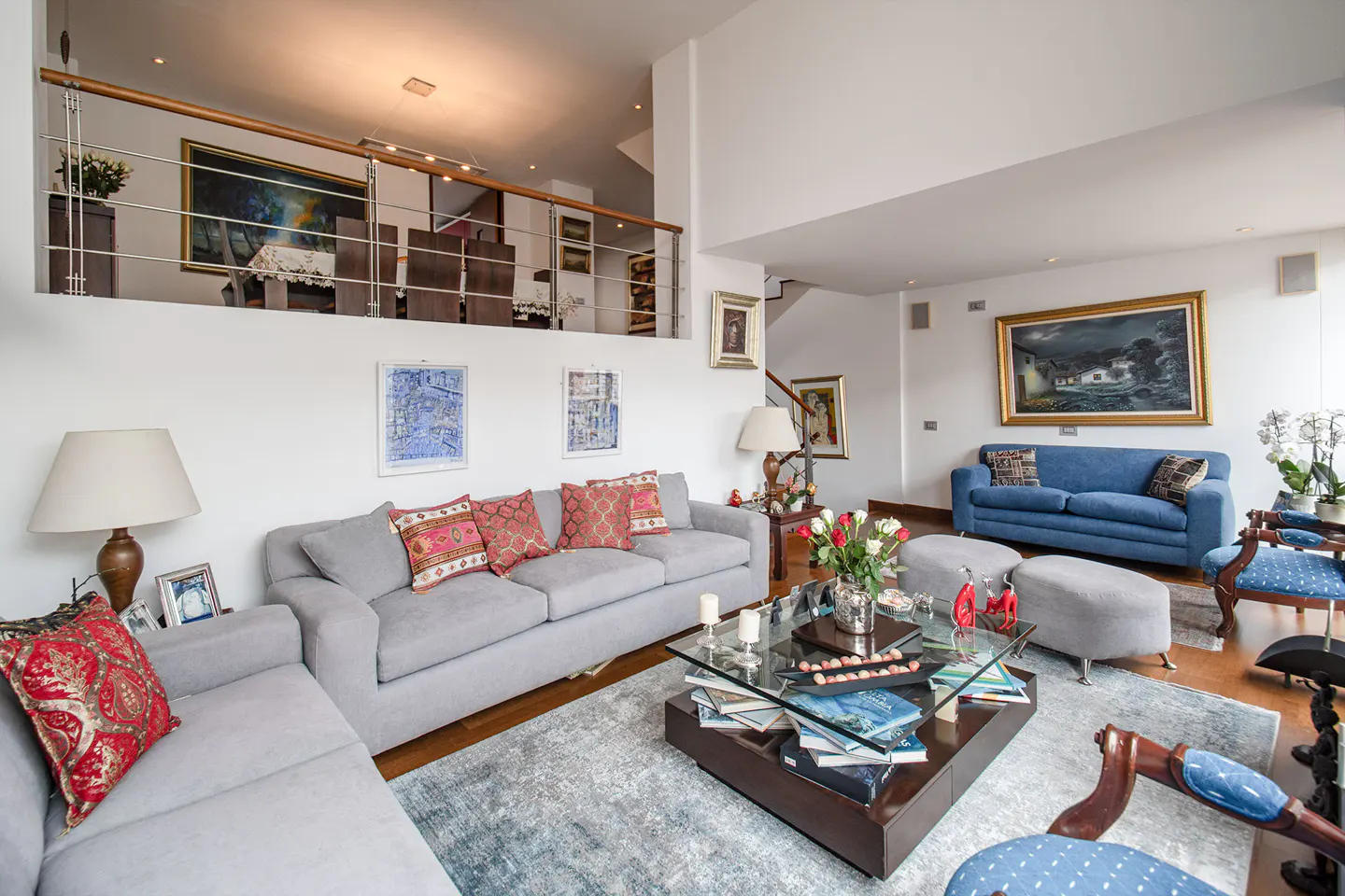 Bright living room with gray sofas, red pillows, and a glass coffee table. A blue sofa sits against the wall with framed art.