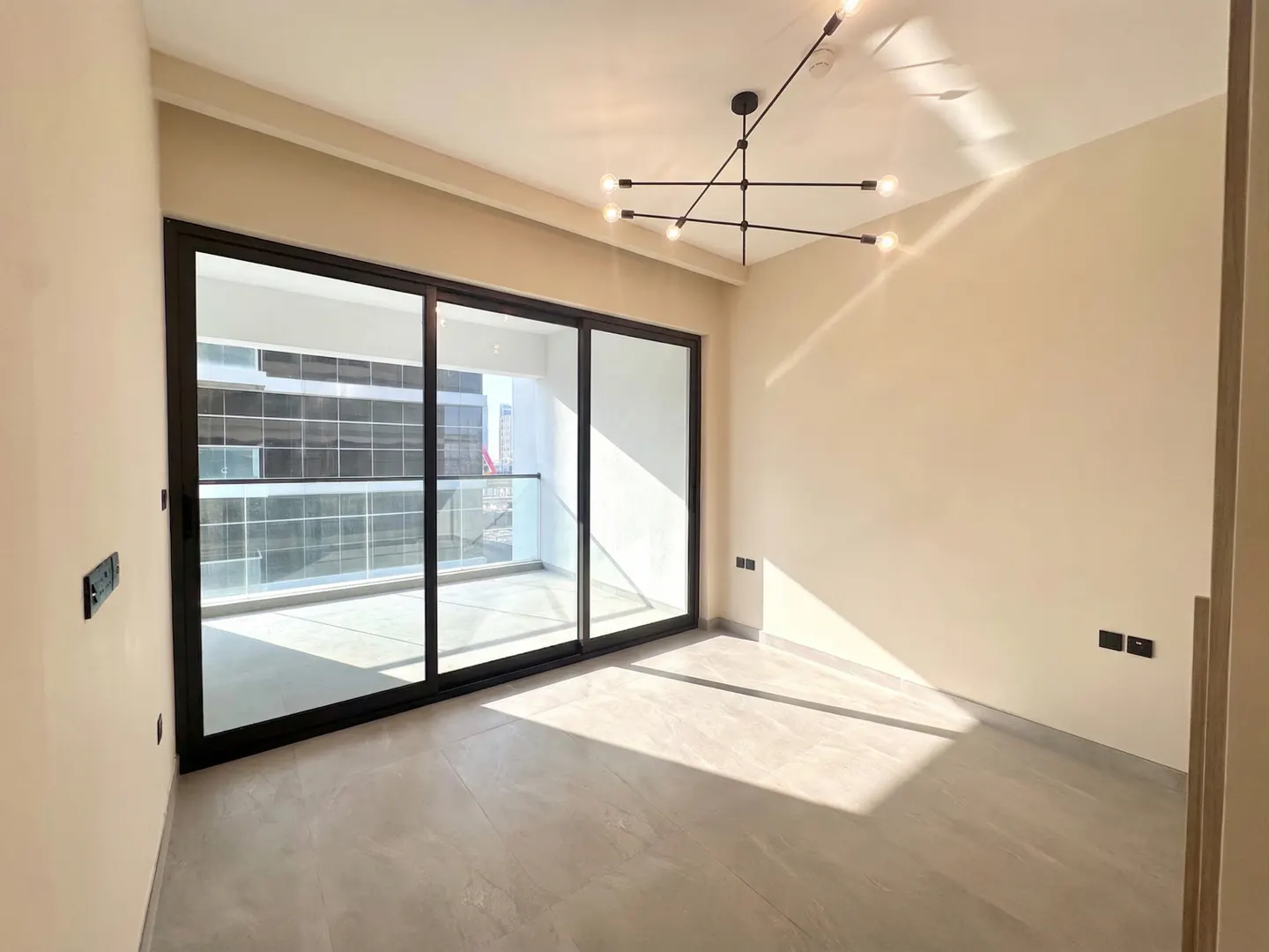 A bright, empty room with beige walls, gray tile floor, and a black-framed sliding glass door to a balcony. A modern black chandelier hangs from the ceiling.