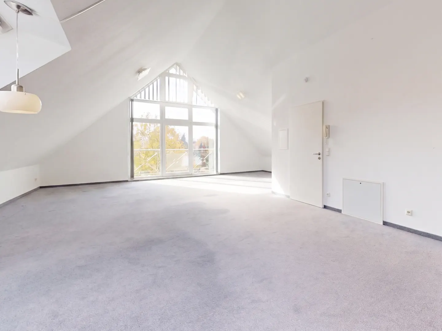Bright, empty attic room with white walls, gray carpet, and a large A-frame window showing trees outside. A white door is on the right.