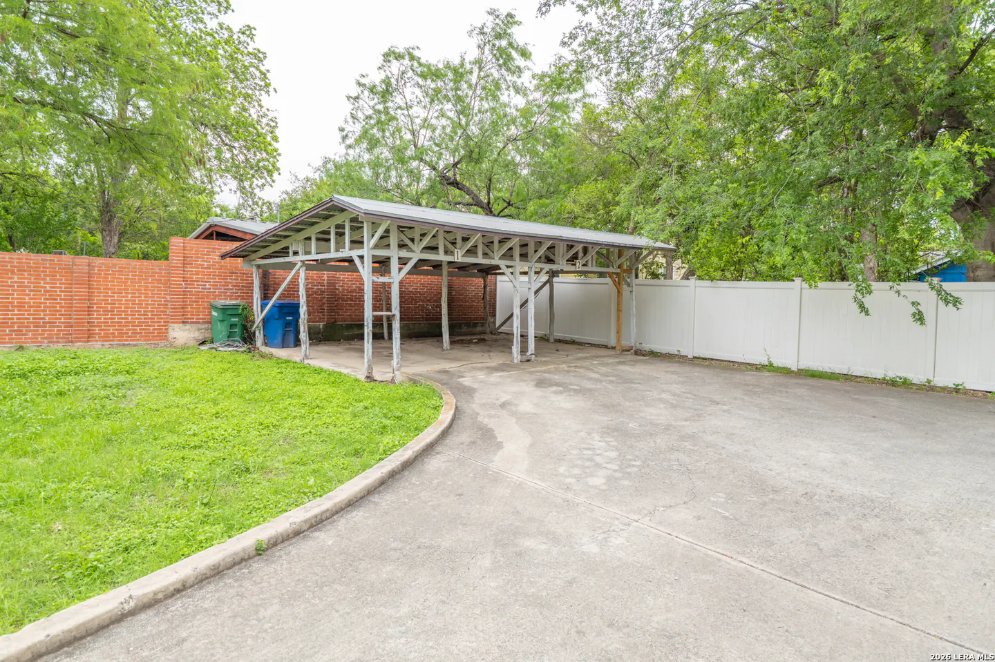 A concrete driveway leads to a white carport, with a brick wall and white fence in the background. Green grass and trees surround the area.