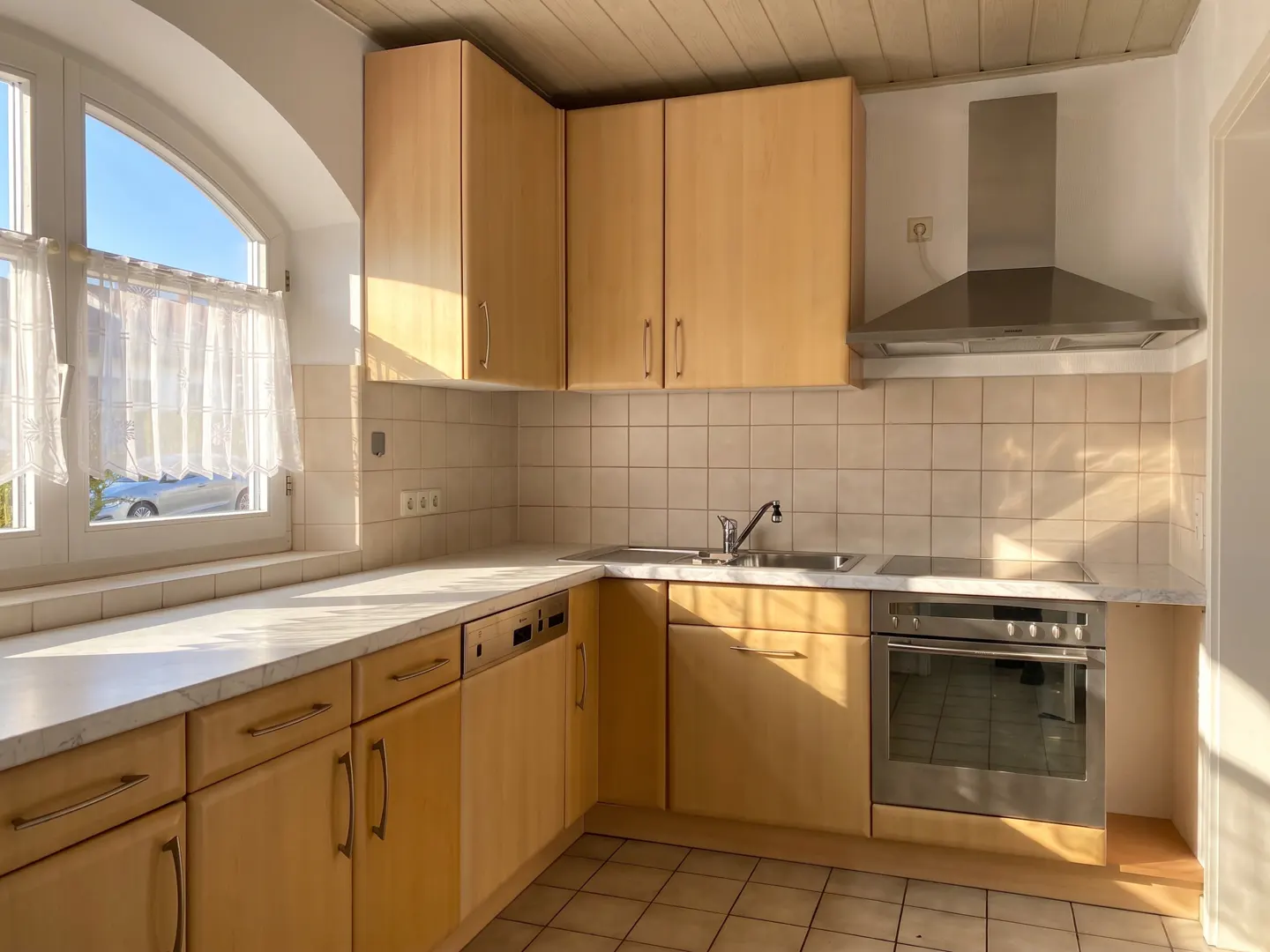 Bright kitchen with light wood cabinets, stainless steel appliances, and tile backsplash. A window with sheer curtains lets in natural light.