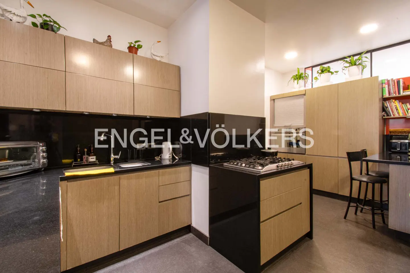 A modern kitchen with light wood cabinets, black countertops, and stainless steel appliances. Plants decorate the top of the cabinets.