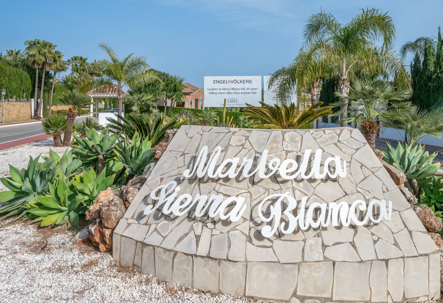 Stone sign reading "Marbella Sierra Blanca" with palm trees and an Engel & Völkers sign in the background under a clear blue sky.