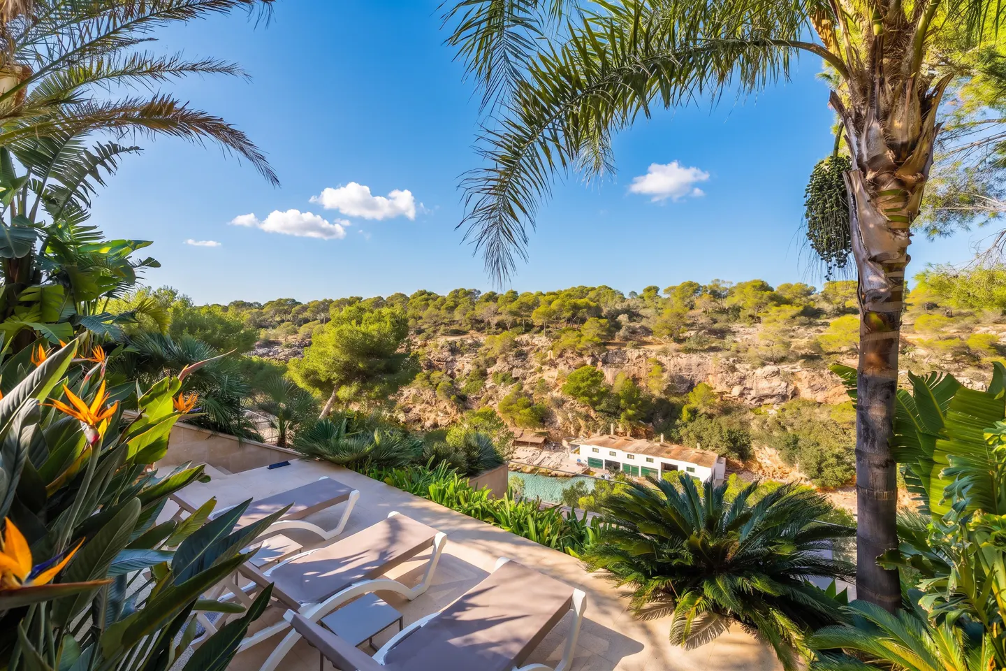 Scenic view of a patio with lounge chairs, lush greenery, and a building nestled in a green hillside under a blue sky.