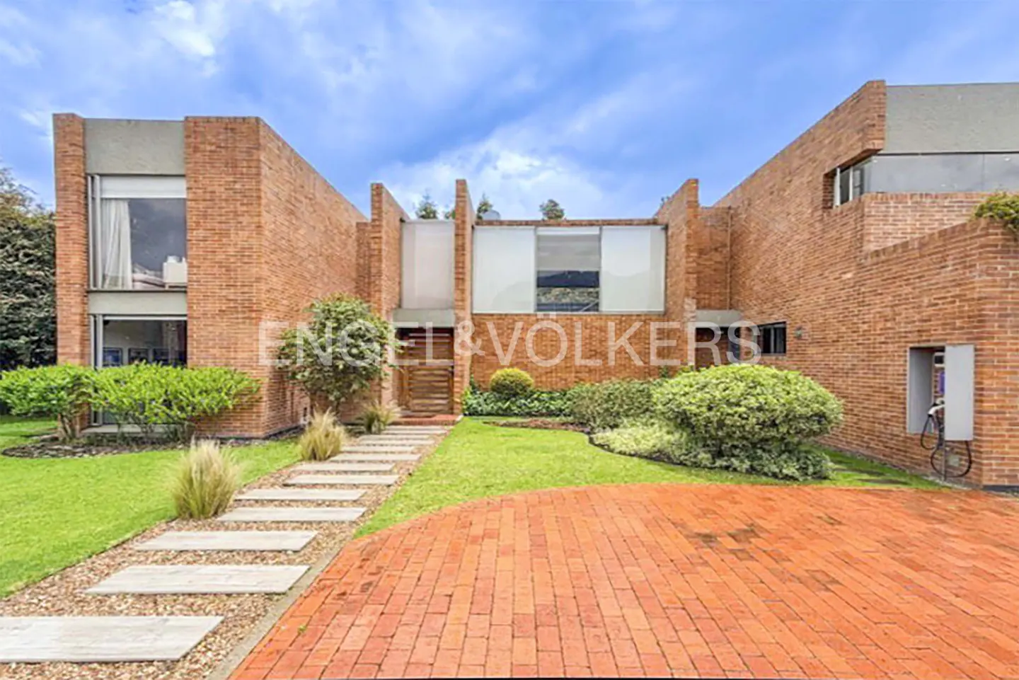 Brick house with a stone walkway leading to the front door, green lawn, and an Engel & Volkers sign.