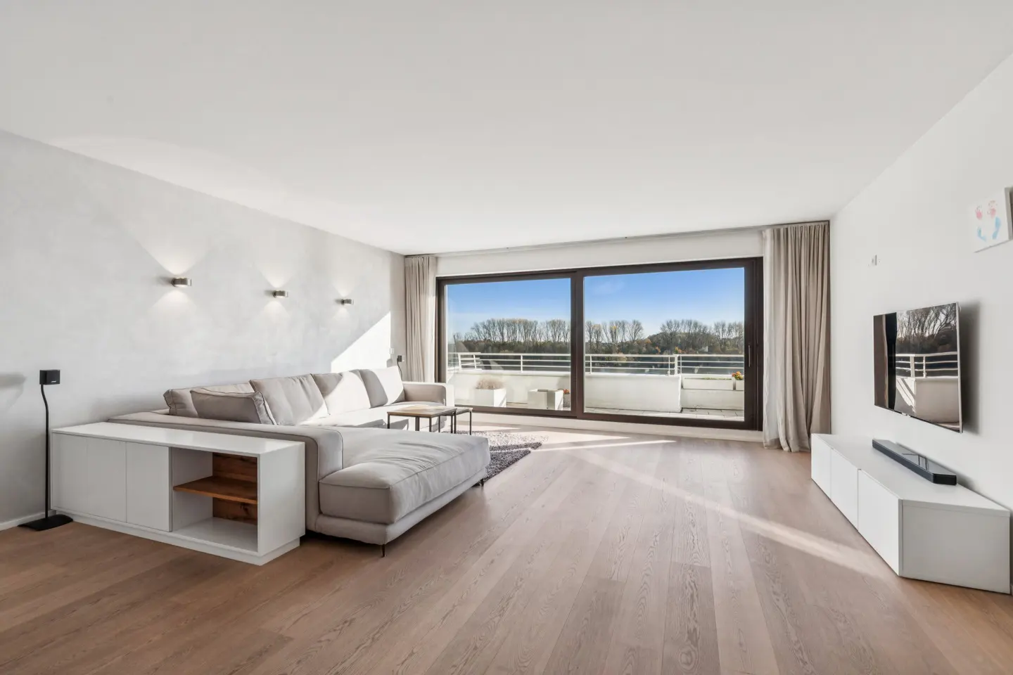 Bright living room with a gray sectional sofa, white cabinets, and a large window overlooking a balcony and trees.