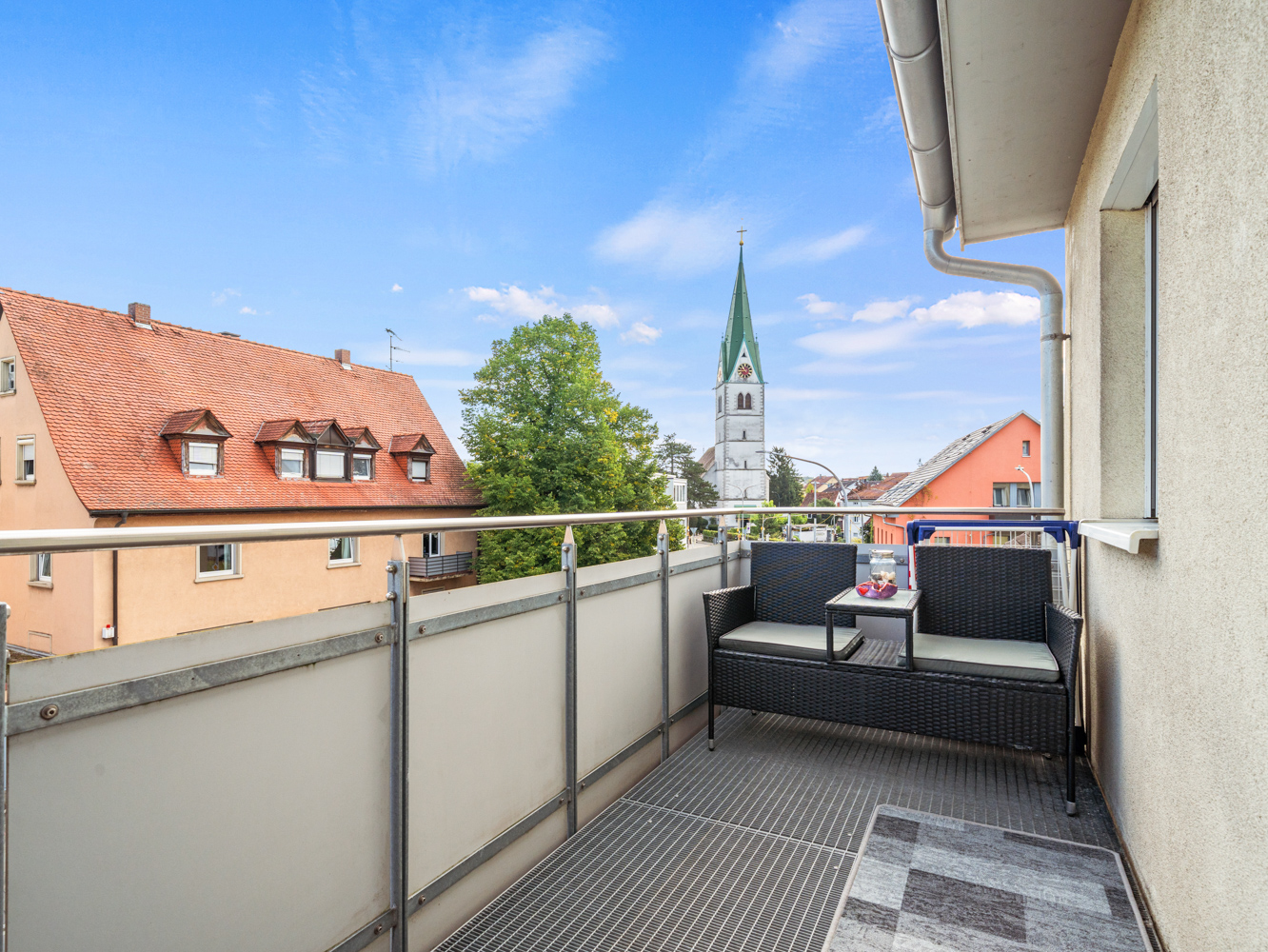 Balcony with black wicker bench and small table, overlooking a town with a church steeple under a bright blue sky. [AI-generated text]