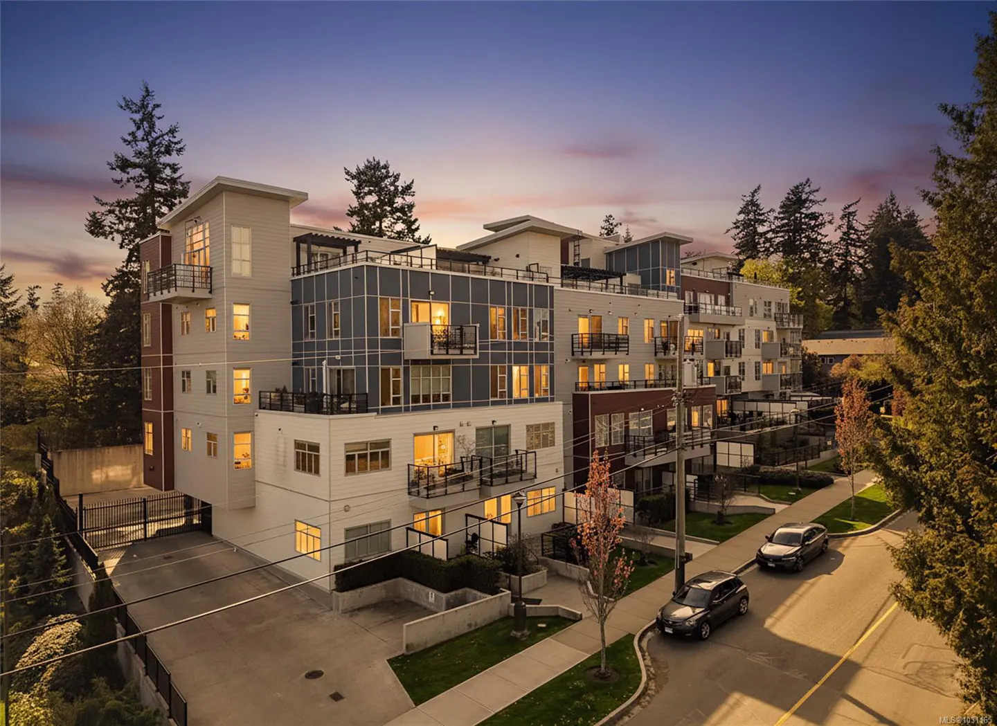 Exterior view of a modern, multi-story apartment building with balconies, cars parked on the street, and trees at dusk.