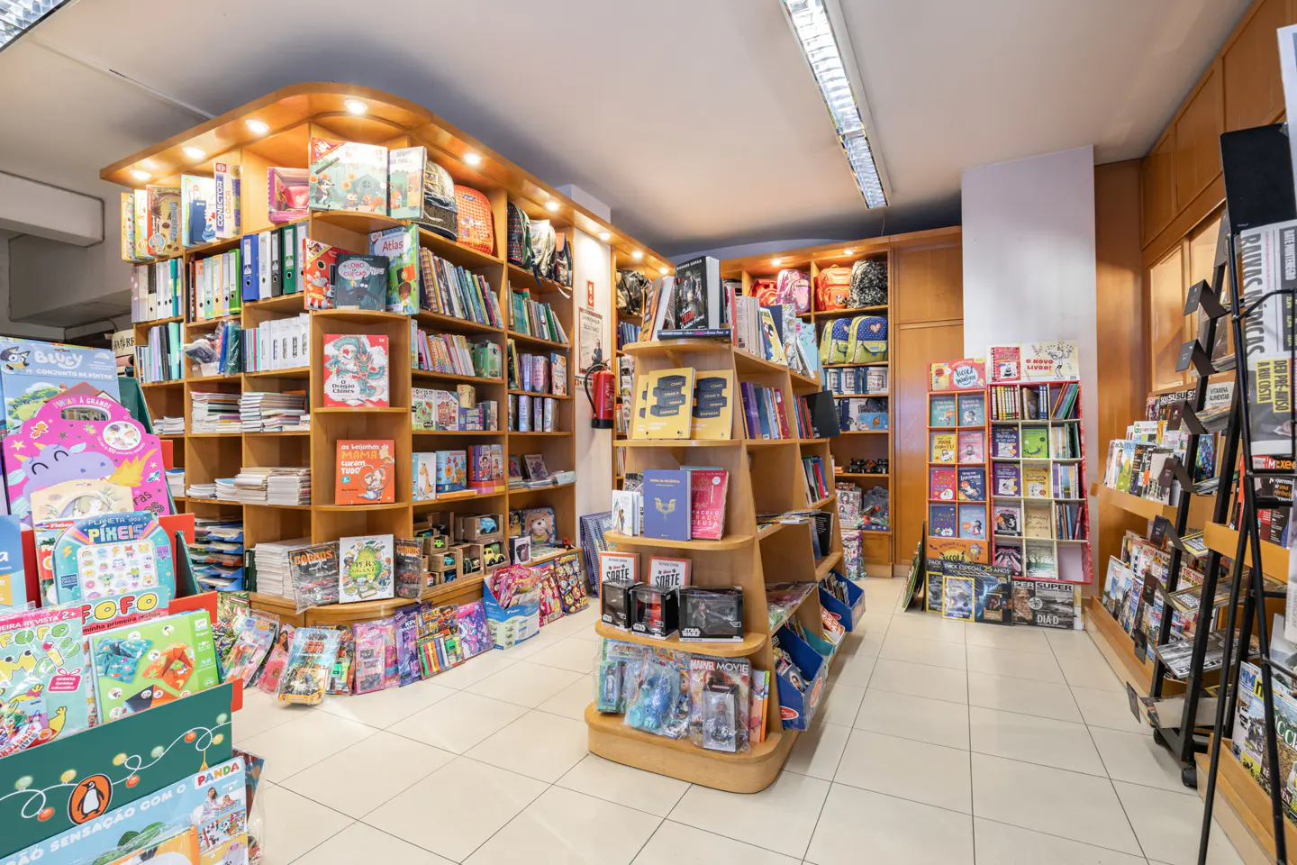 Interior view of a bookstore with wooden shelves filled with colorful books and toys on a white tiled floor.