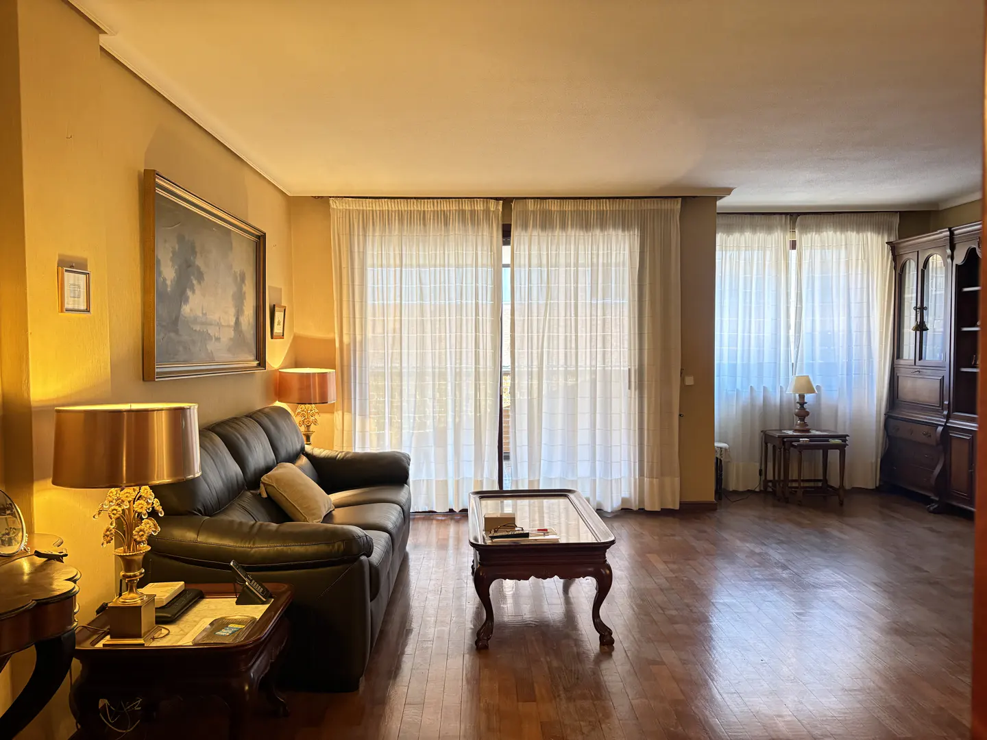 Living room with dark leather sofa, wood floors, coffee table, and sheer white curtains. A painting hangs on the yellow wall.