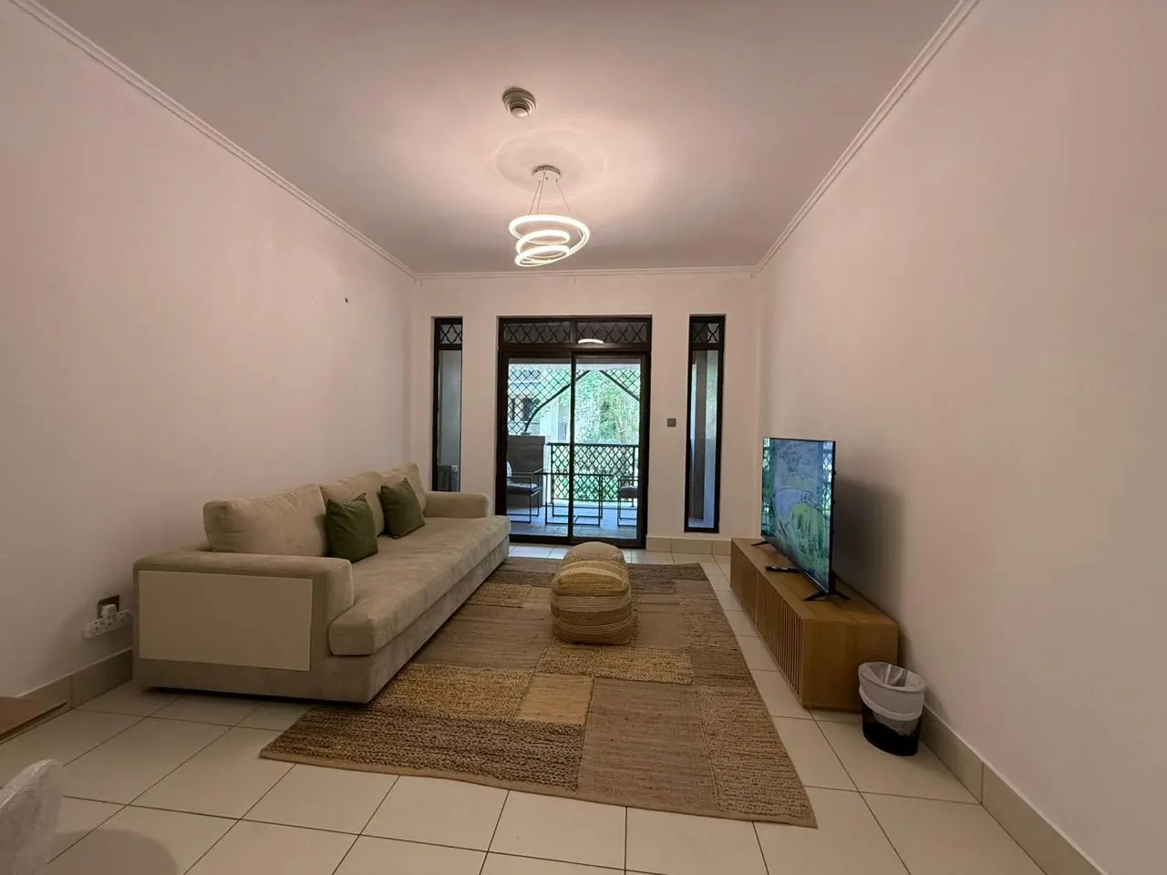 Living room with beige sofa, green pillows, and a brown patchwork rug. Sliding glass doors lead to a balcony. A TV sits on a wooden console.