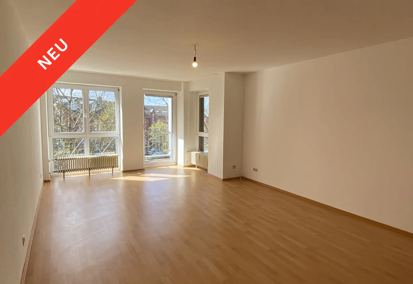 Bright, empty room with wood floors, white walls, and large windows. A red "NEU" banner is in the upper left corner.