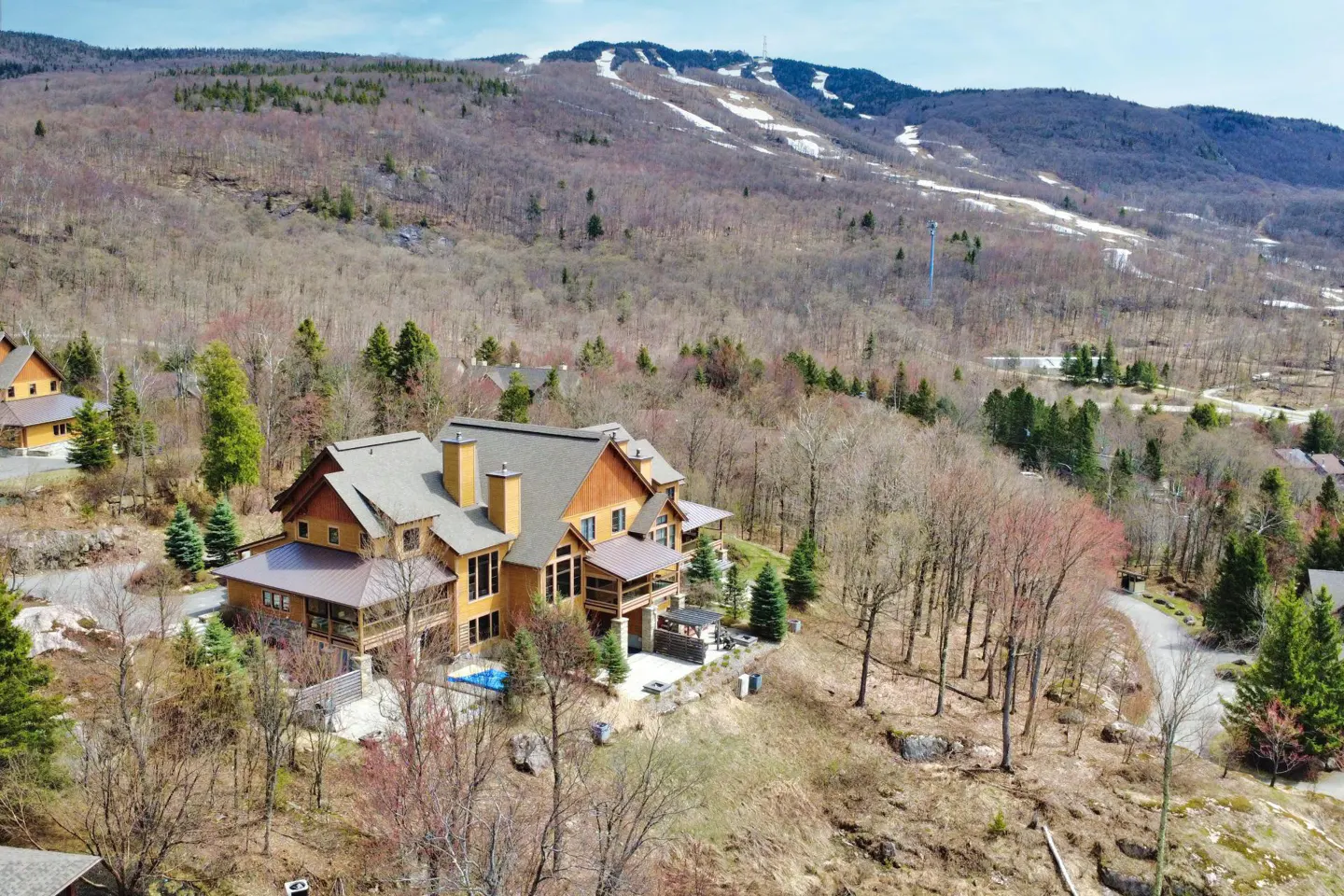 Aerial view of a large, multi-story wooden mountain home nestled in a forest with a ski slope visible on the mountain behind.