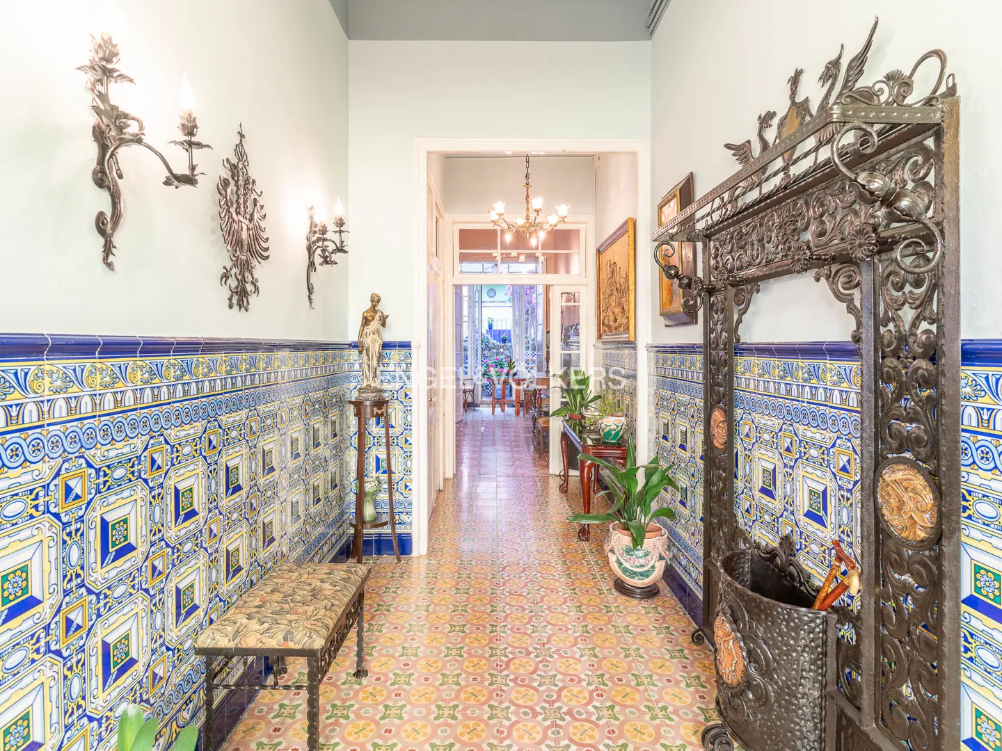 Hallway with blue and yellow tiled walls and floor, ornate metal coat rack, sconces, and a statue.