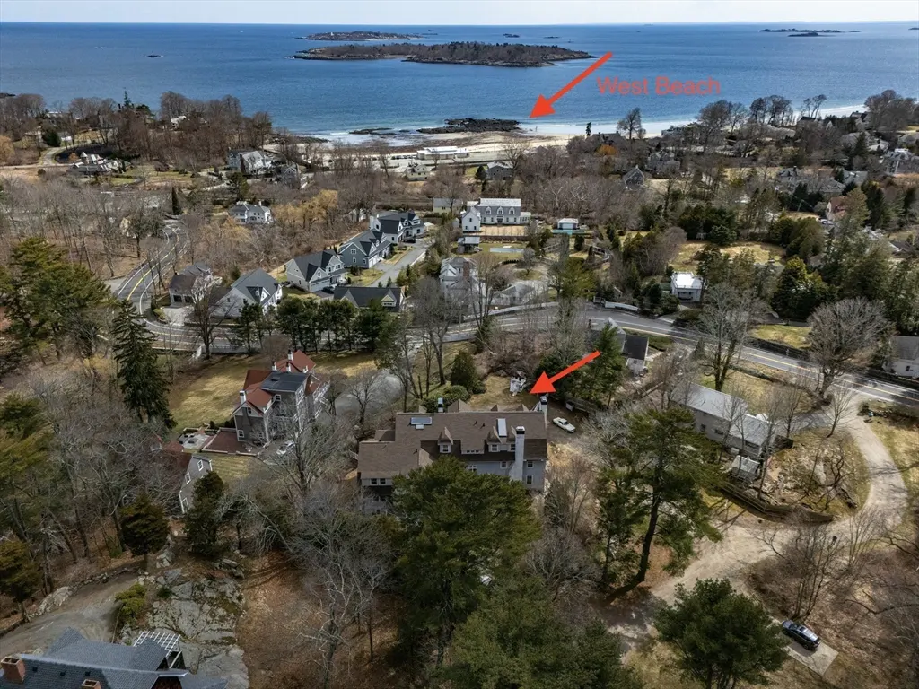 Aerial view of a neighborhood with houses, trees, and a beach in the background. The sky is blue and the water is calm.