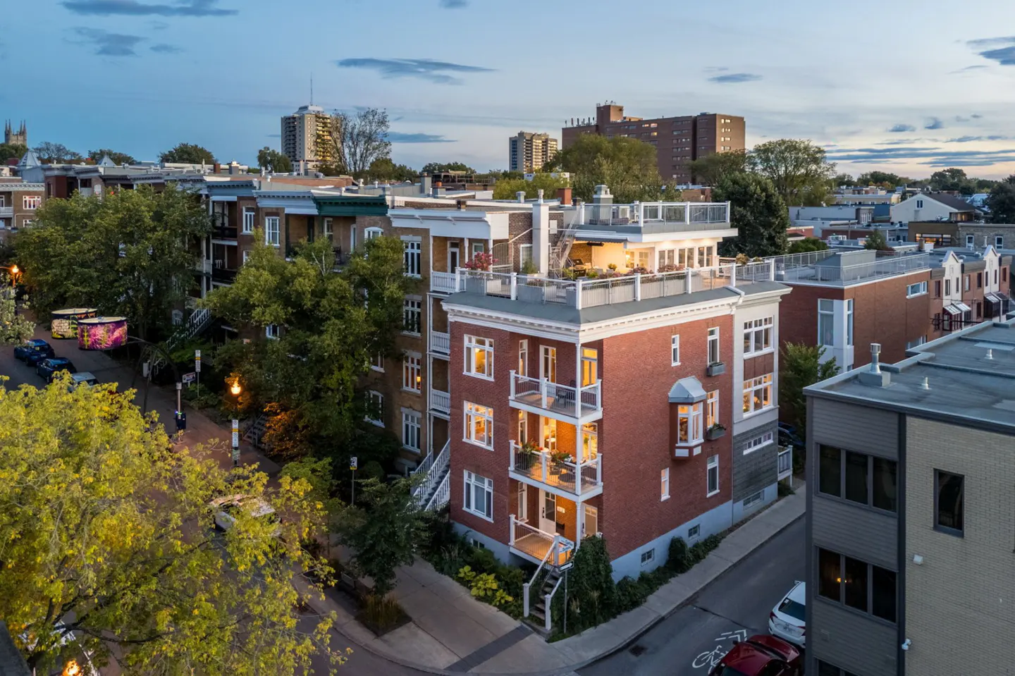 Aerial view of a red brick apartment building with lit balconies and a rooftop terrace in a city neighborhood at dusk.