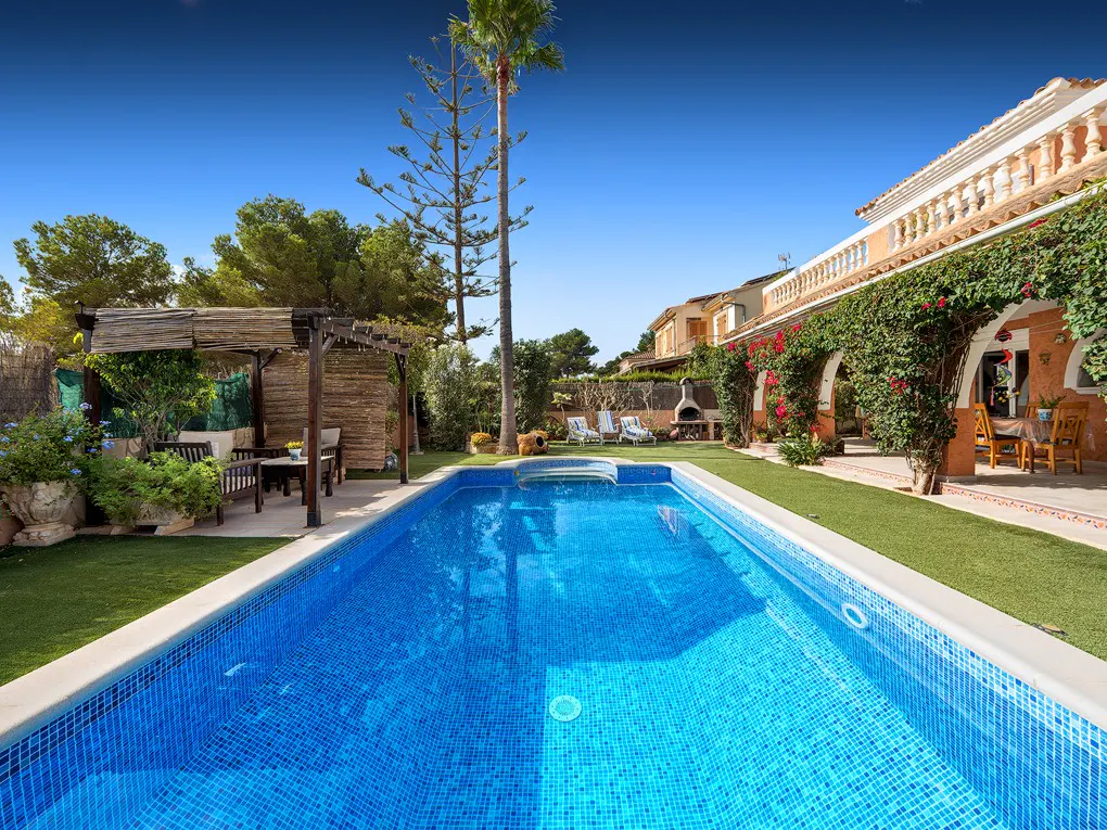 A rectangular blue tiled pool is centered in a green lawn, with a pergola, lounge chairs, and a house with red flowers in the background.