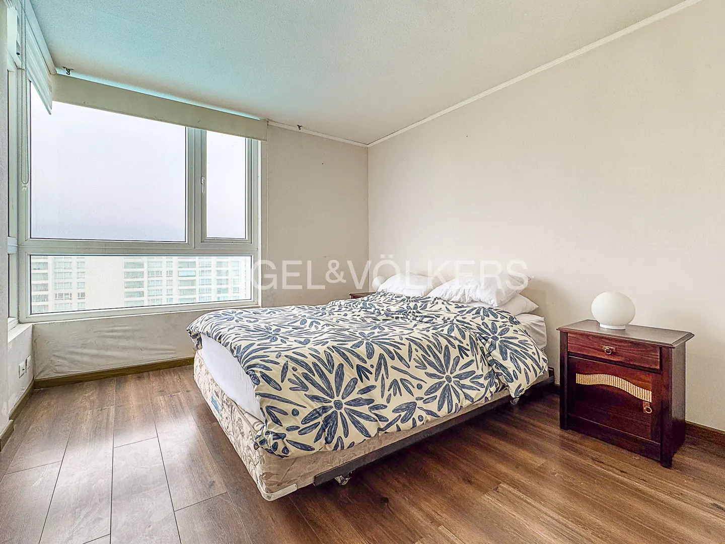 Bedroom with a large window, wood floors, a bed with a blue floral comforter, and a dark wood nightstand.