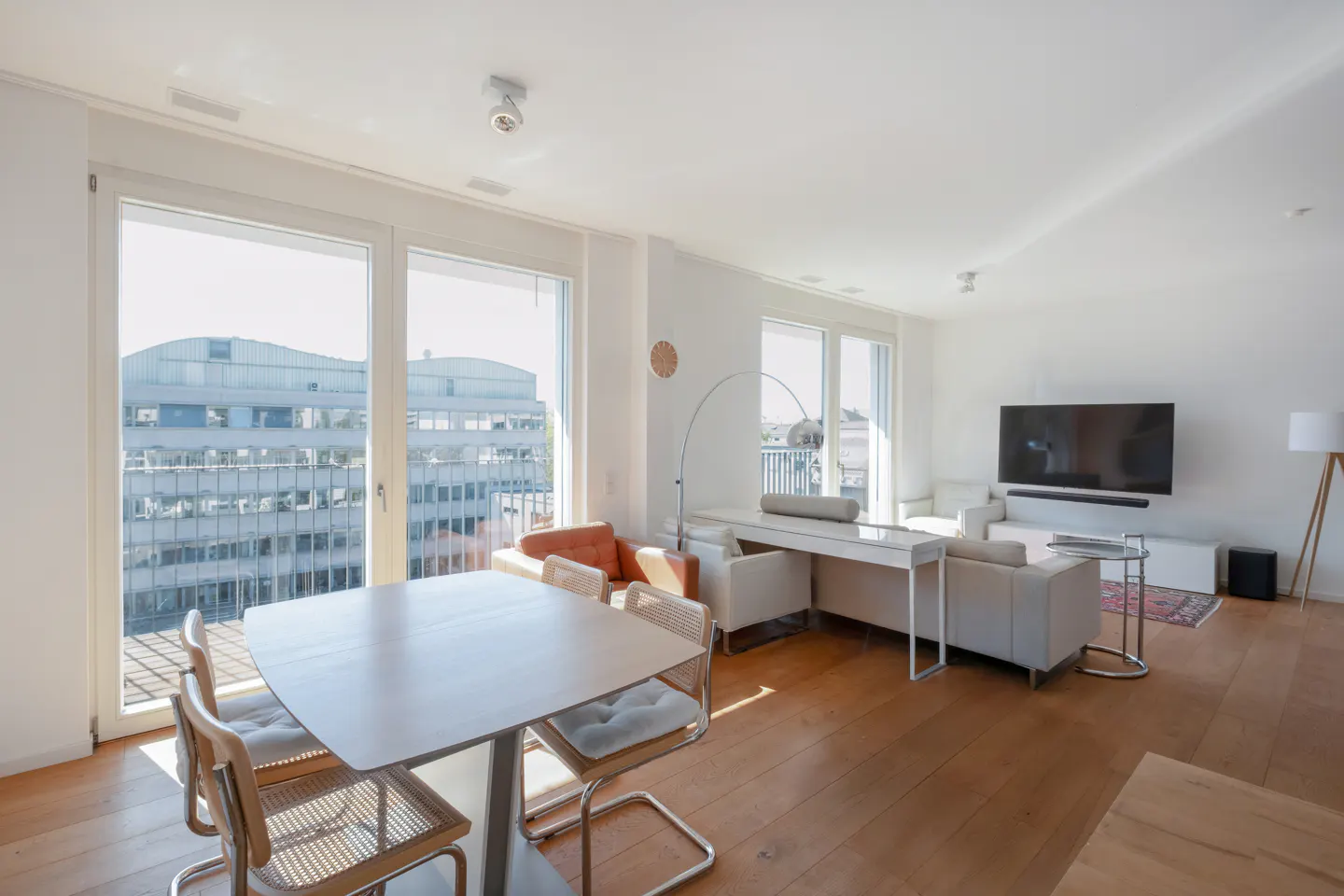 Bright, modern living room with hardwood floors, dining area, white sofa, wall-mounted TV, and large windows with city view.