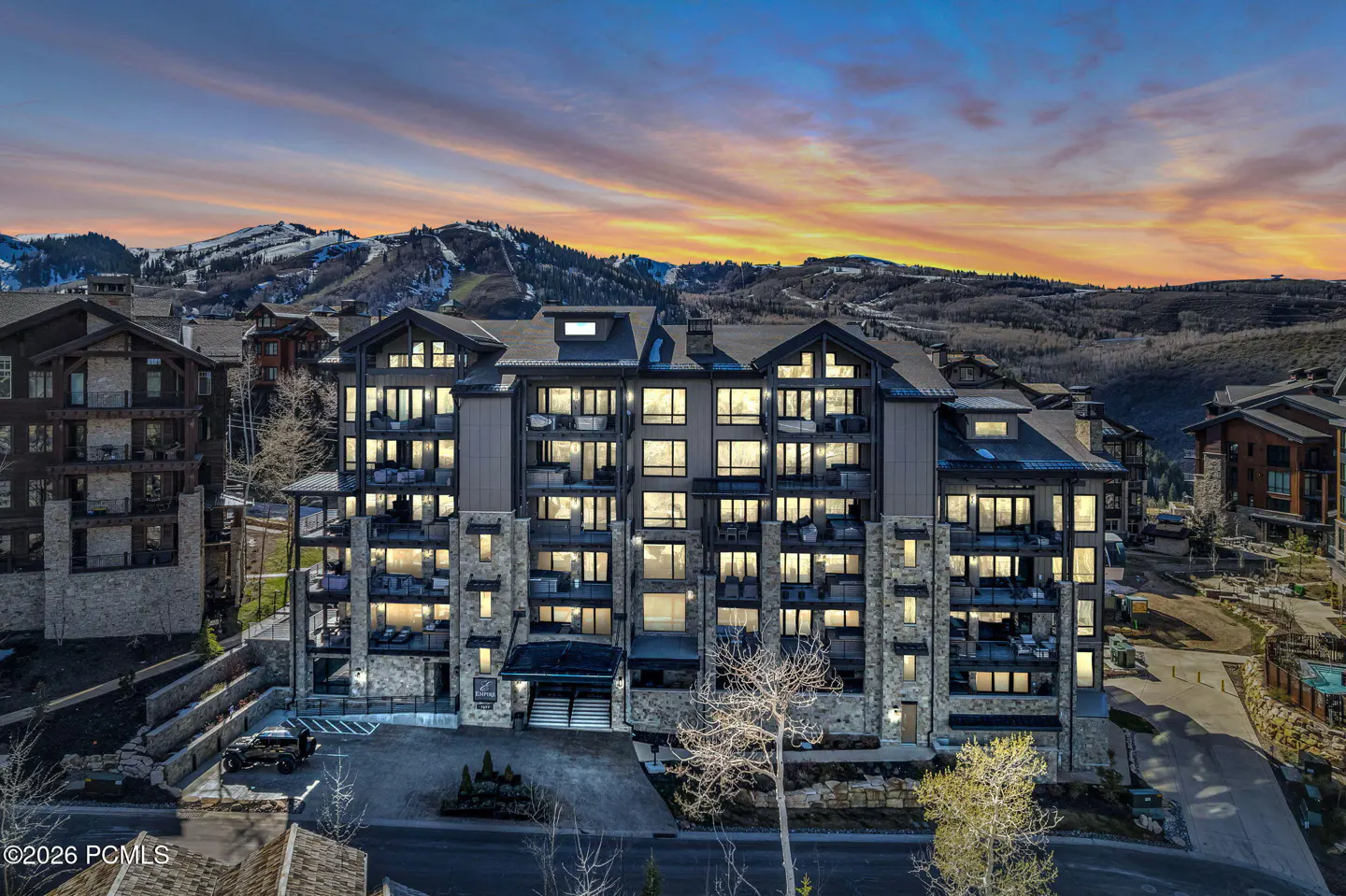 Exterior view of a stone and gray condo building with lit windows at dusk, mountains in the background.