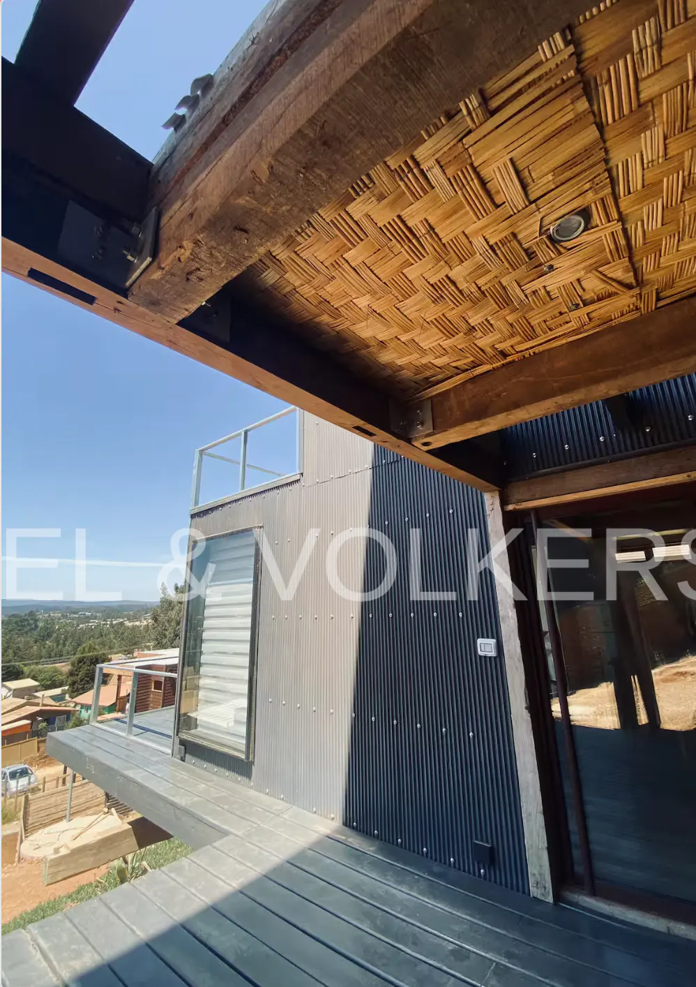 Modern home exterior with a woven bamboo ceiling, corrugated metal walls, and a wooden deck overlooking a rural landscape.