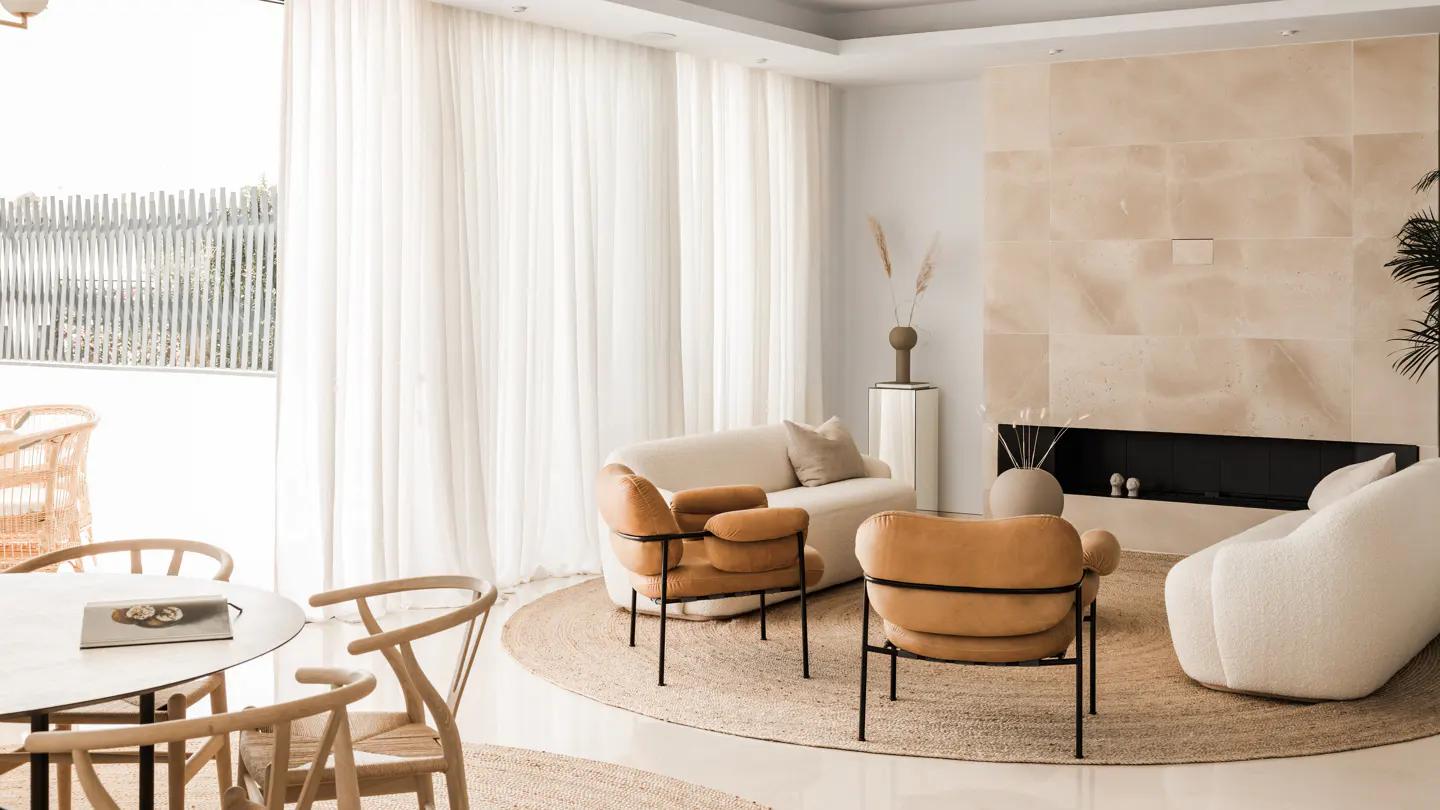 Bright living room with beige sofas, tan chairs, and a stone fireplace. Sheer white curtains cover a large window.