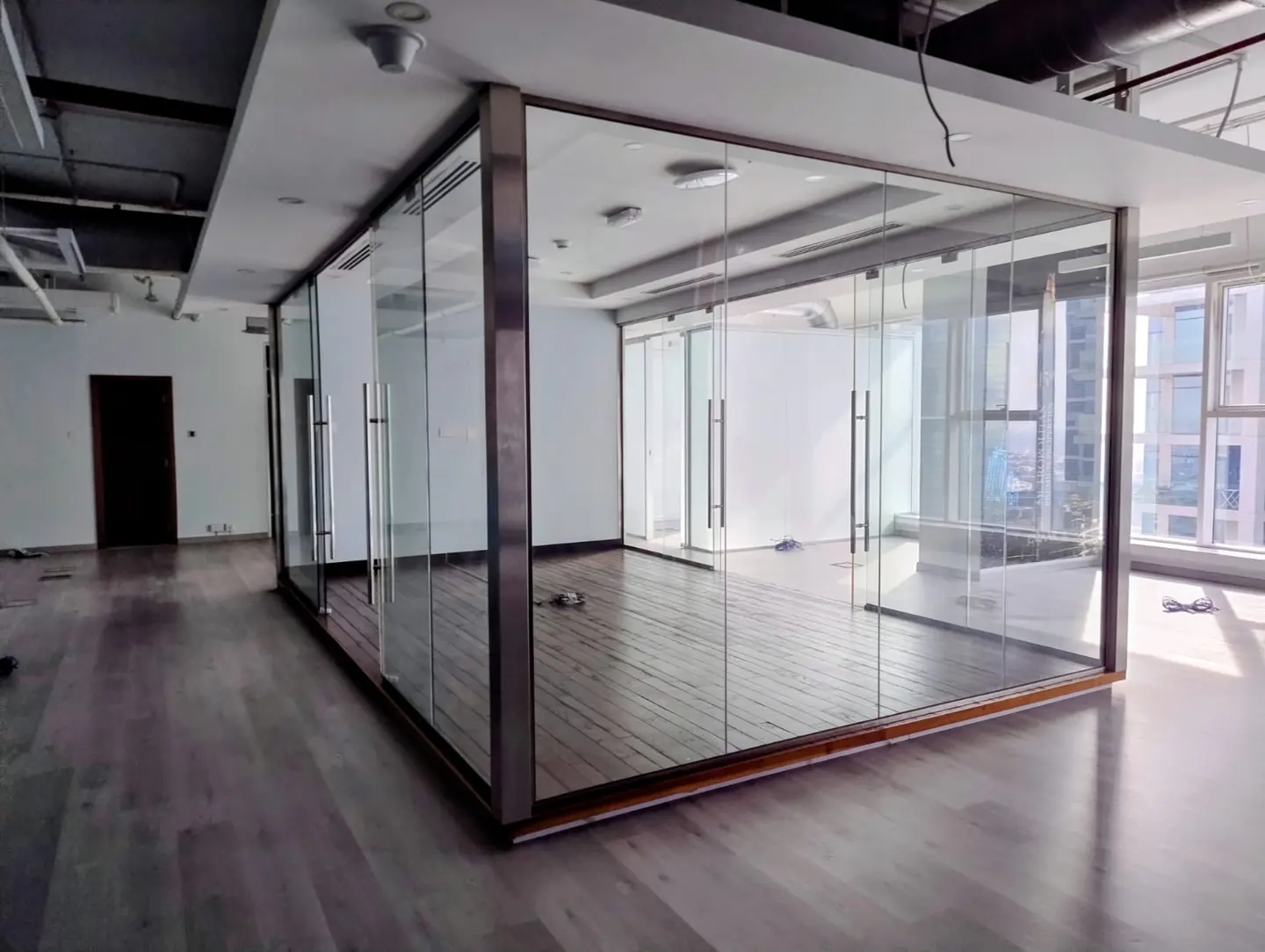Empty office space with glass-walled conference room. Wood floors, white walls, and large windows with city views.