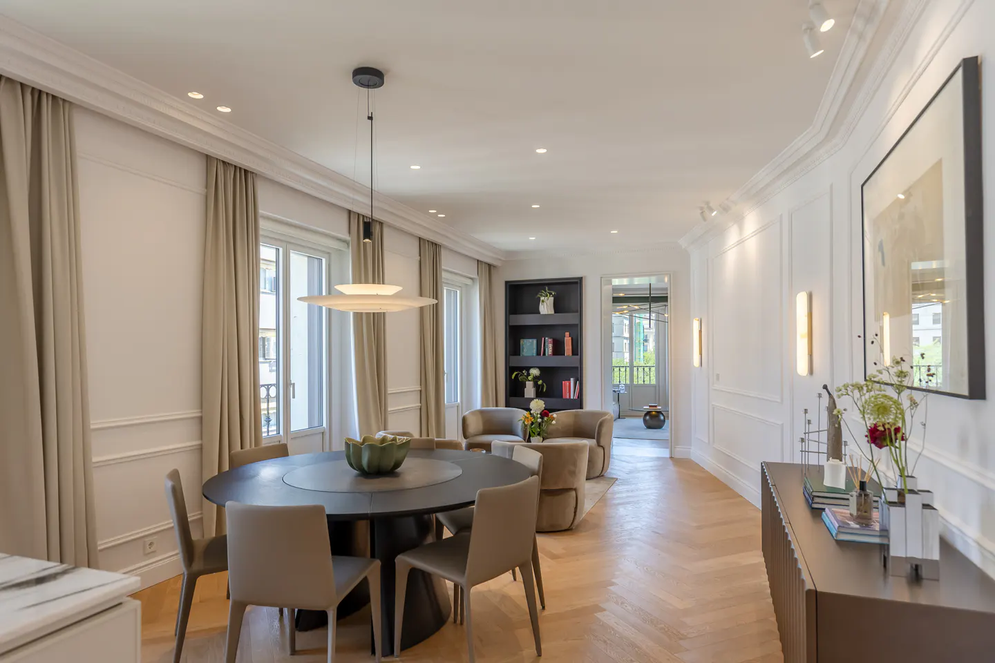 Elegant dining room with a round black table, beige chairs, herringbone wood floors, and a view into a cozy lounge area.