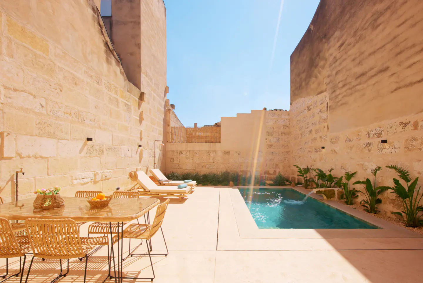 Outdoor patio with a pool, table, and lounge chairs. The patio is surrounded by stone walls and the sky is blue.