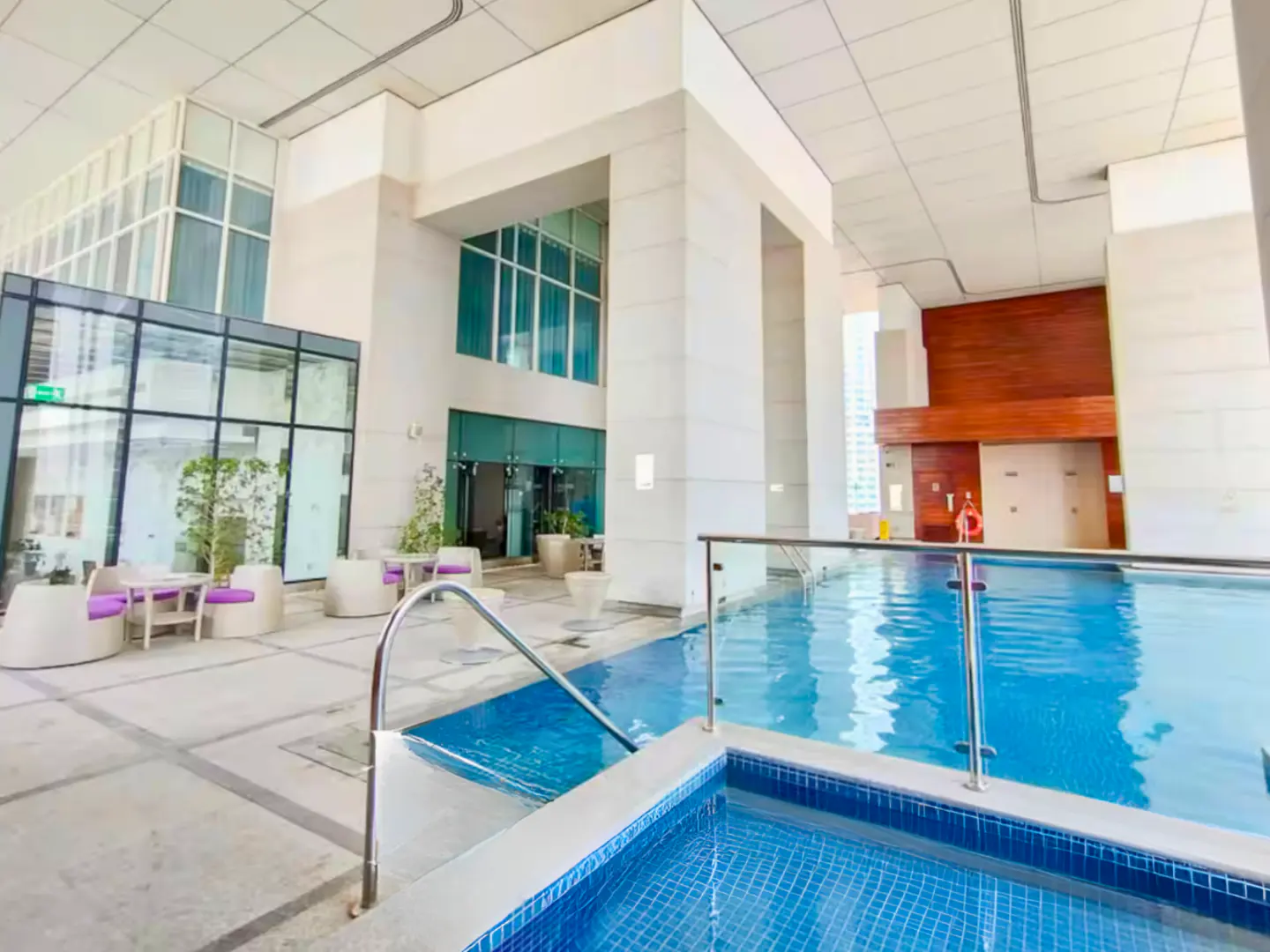 Indoor pool with blue mosaic tiles and stainless steel handrails. Lounge area with white chairs and purple cushions. Large windows and white pillars.