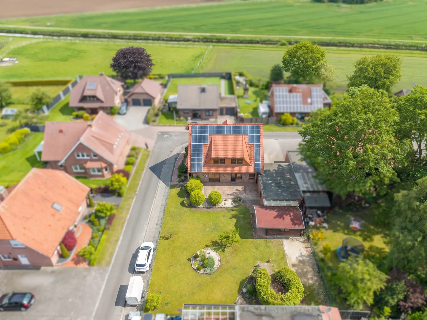 Aerial view of a red-roofed house with solar panels, surrounded by green lawns, trees, and other houses in a rural neighborhood.