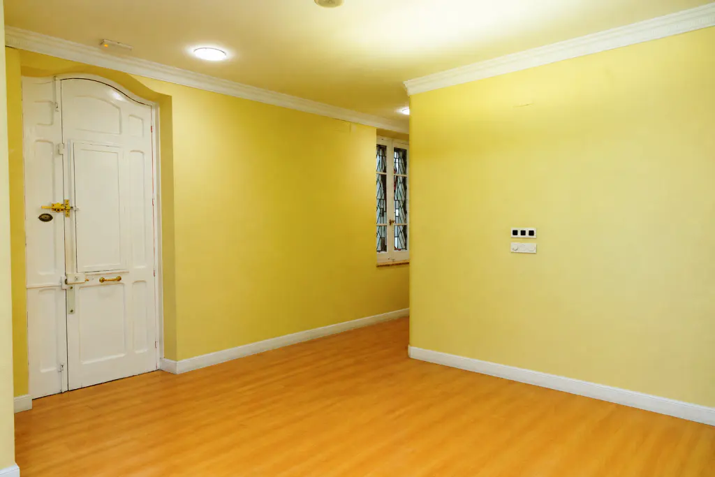 Empty room with yellow walls, white trim, and wood floors. A white door with gold hardware is on the left. A window is visible in the background.