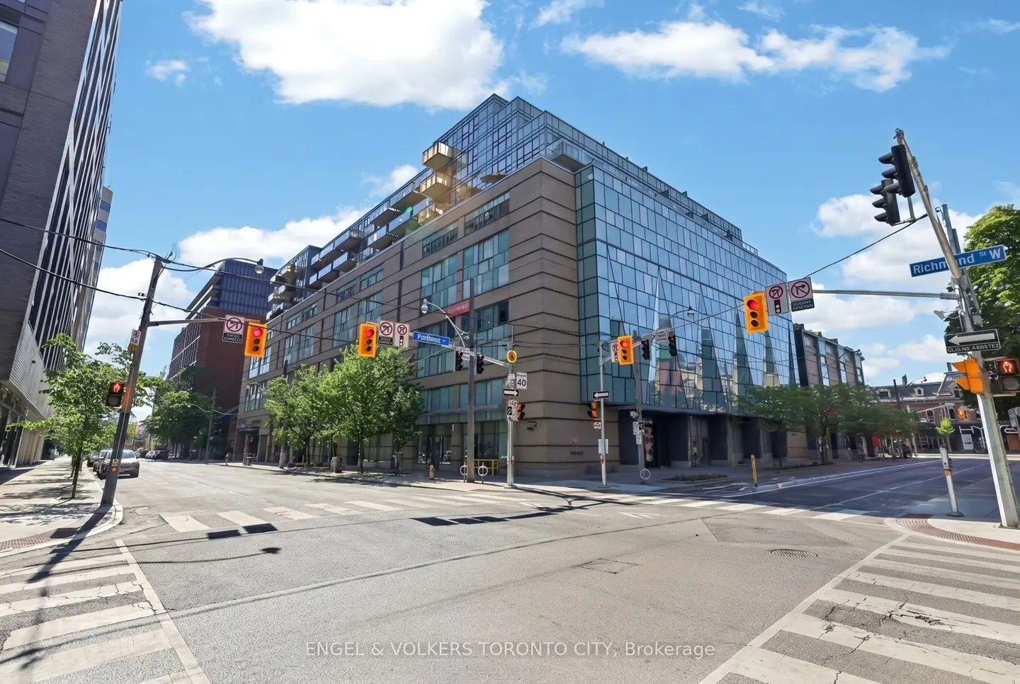 A modern glass and brick mid-rise building on a sunny city street corner with traffic lights and crosswalks.