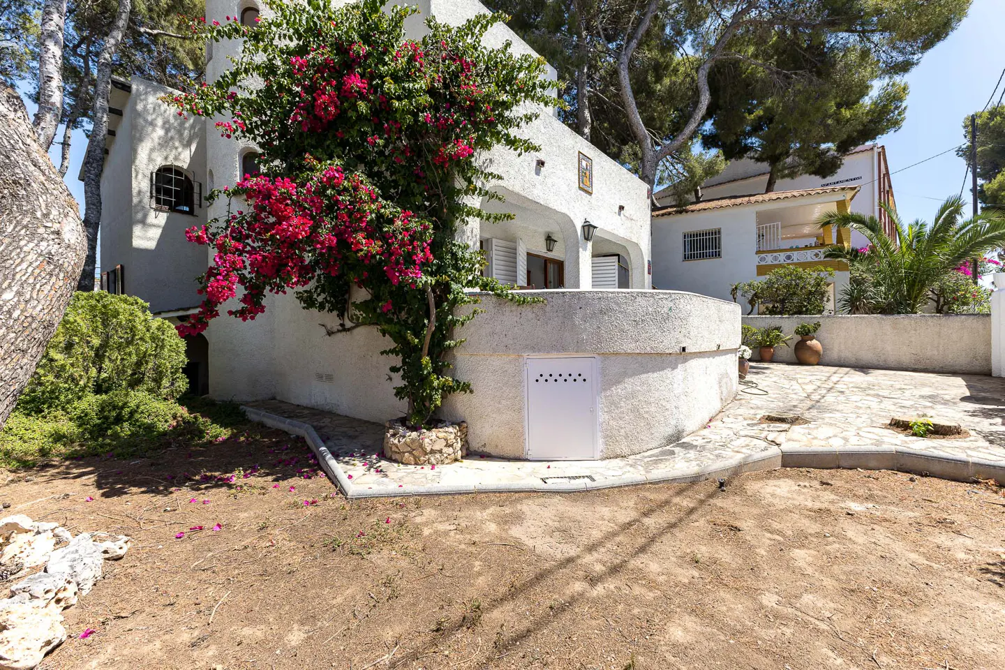 White Mediterranean-style house with a vibrant pink bougainvillea, stone patio, and surrounding trees under a blue sky.