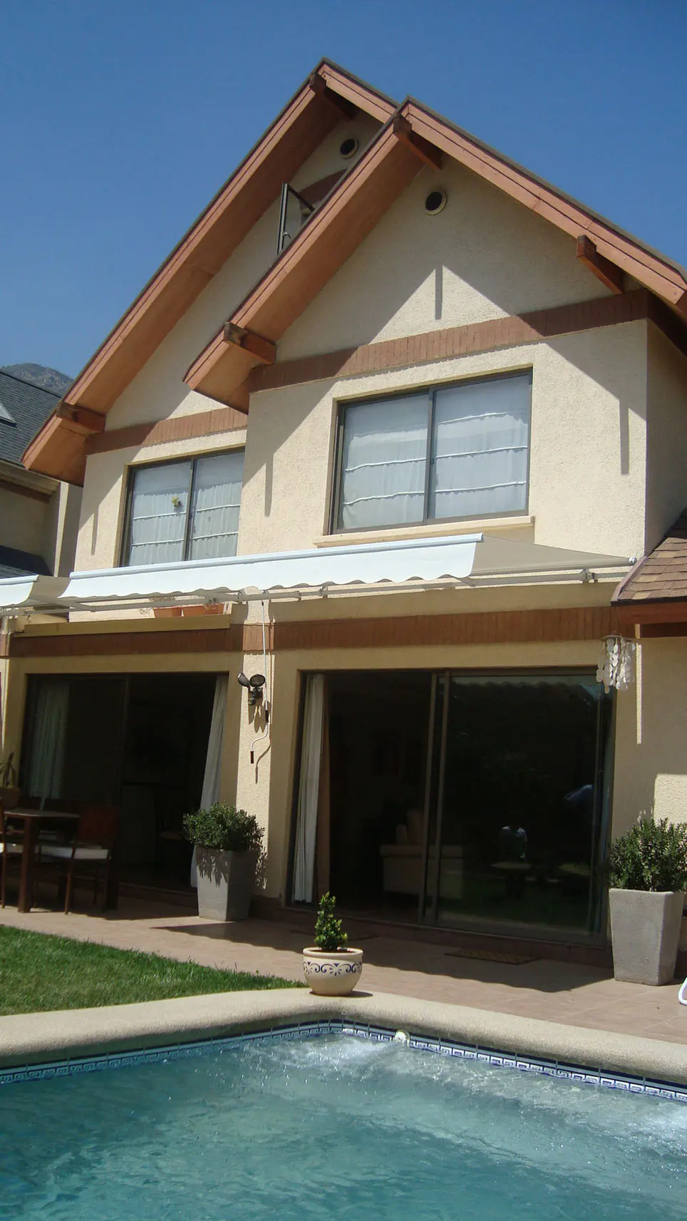 Two-story beige house with brown trim, a white awning, and a blue pool in the foreground. Sliding glass doors open to a patio.