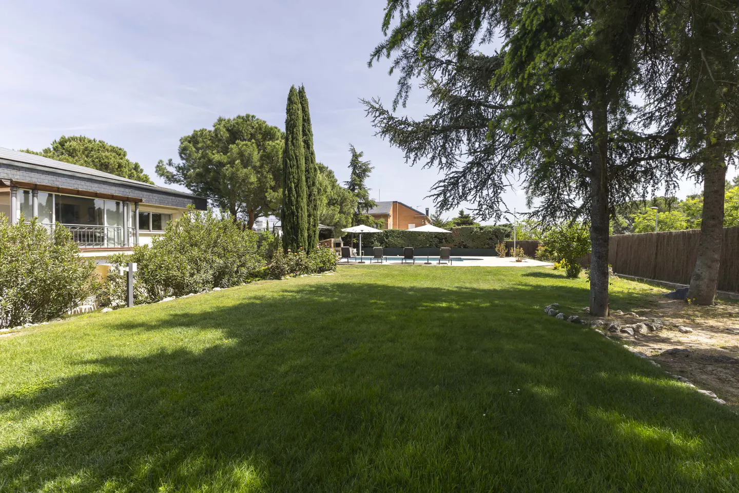 A lush green lawn leads to a pool with lounge chairs and umbrellas, framed by trees and a house in the background.