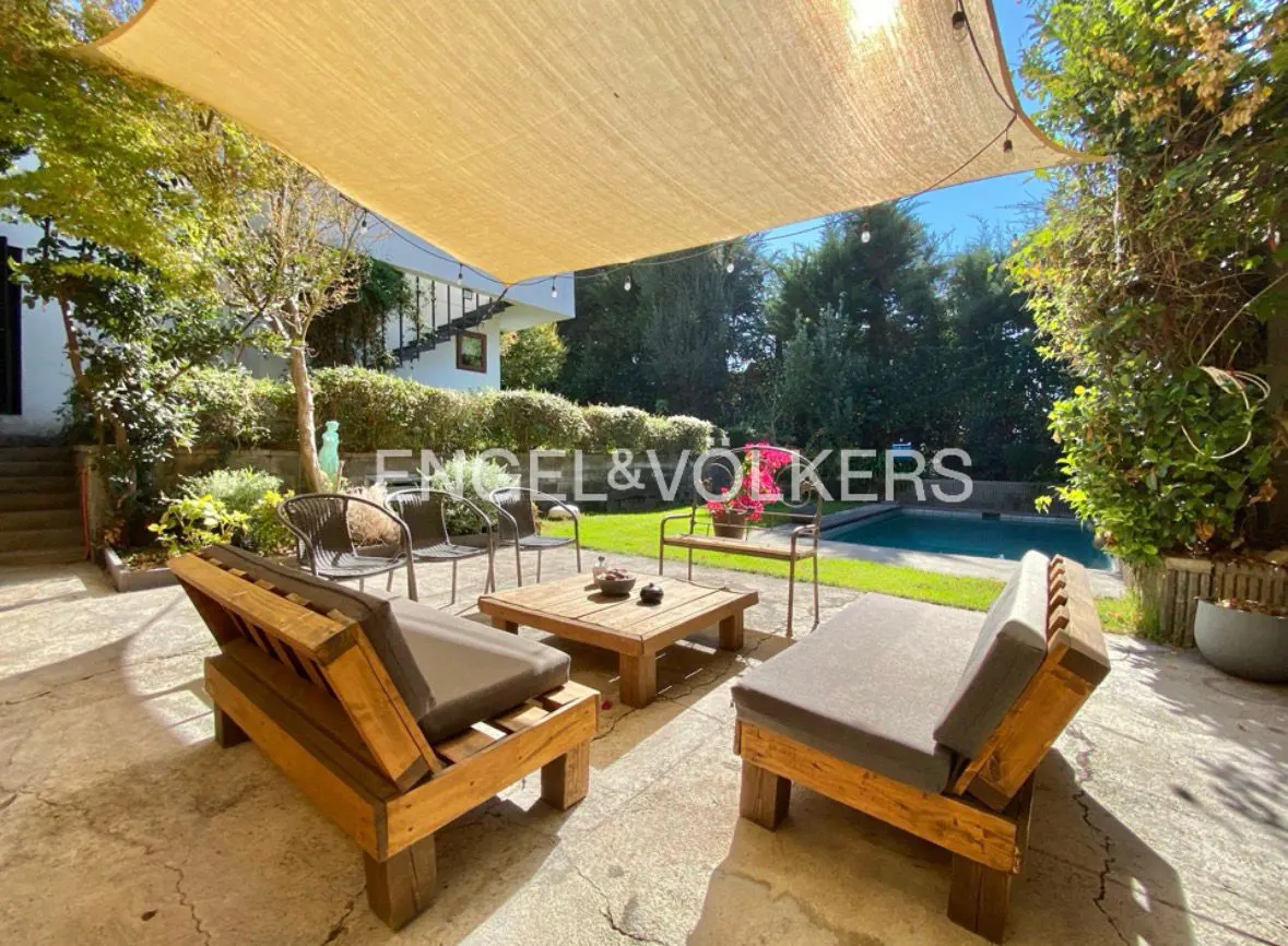 Outdoor patio with wooden furniture, gray cushions, and a beige sunshade. A pool and lush greenery are in the background.