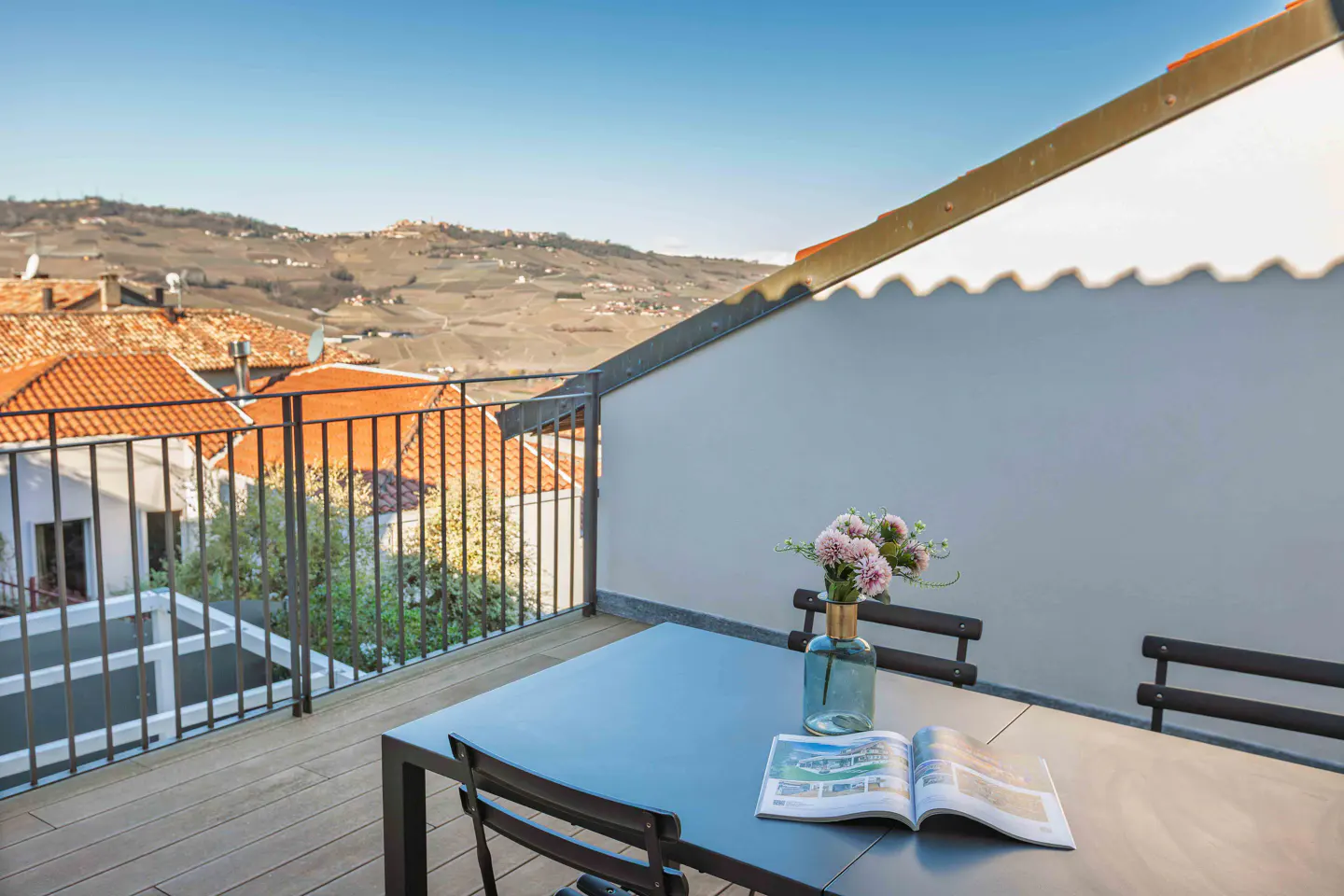 Outdoor patio with a black table, chairs, flowers in a vase, and a magazine. Rolling hills and orange rooftops in the background.