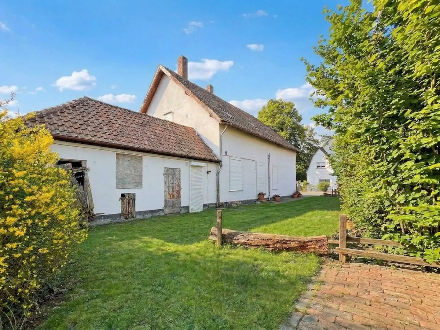 Exterior view of a white, two-story house with a red tile roof, boarded windows, and a green lawn on a sunny day.